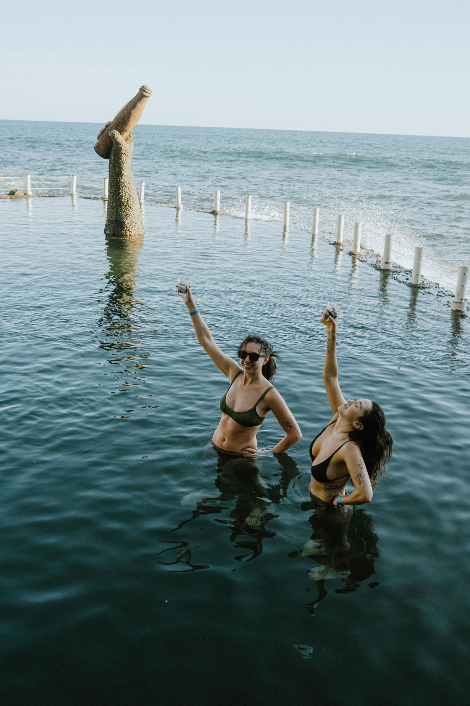 Two swimmers enjoy a unique ocean pool with a tall sculptural post rising from the water.