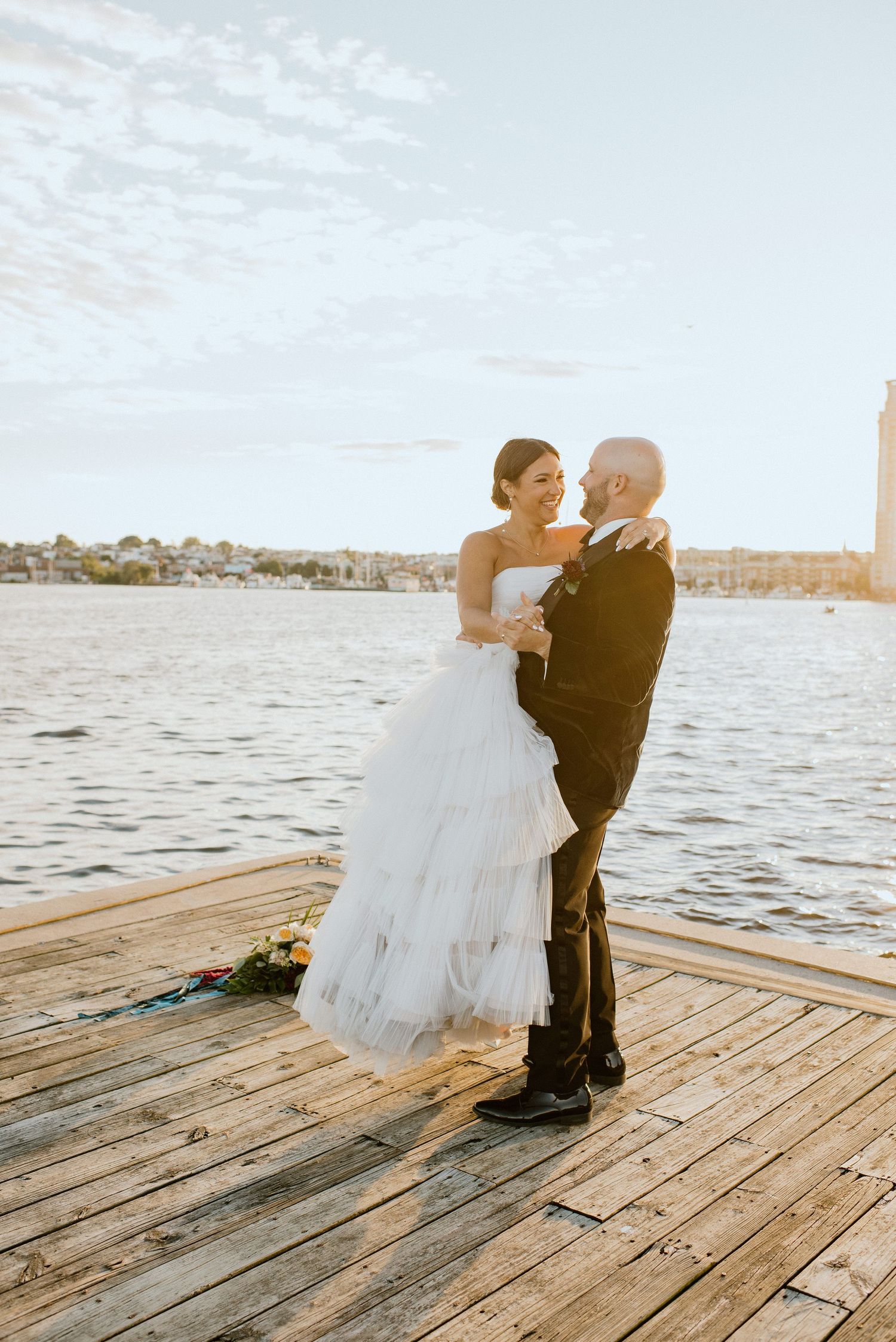 Wedding couple embraces on a waterfront dock during sunset with water and coastline in background.