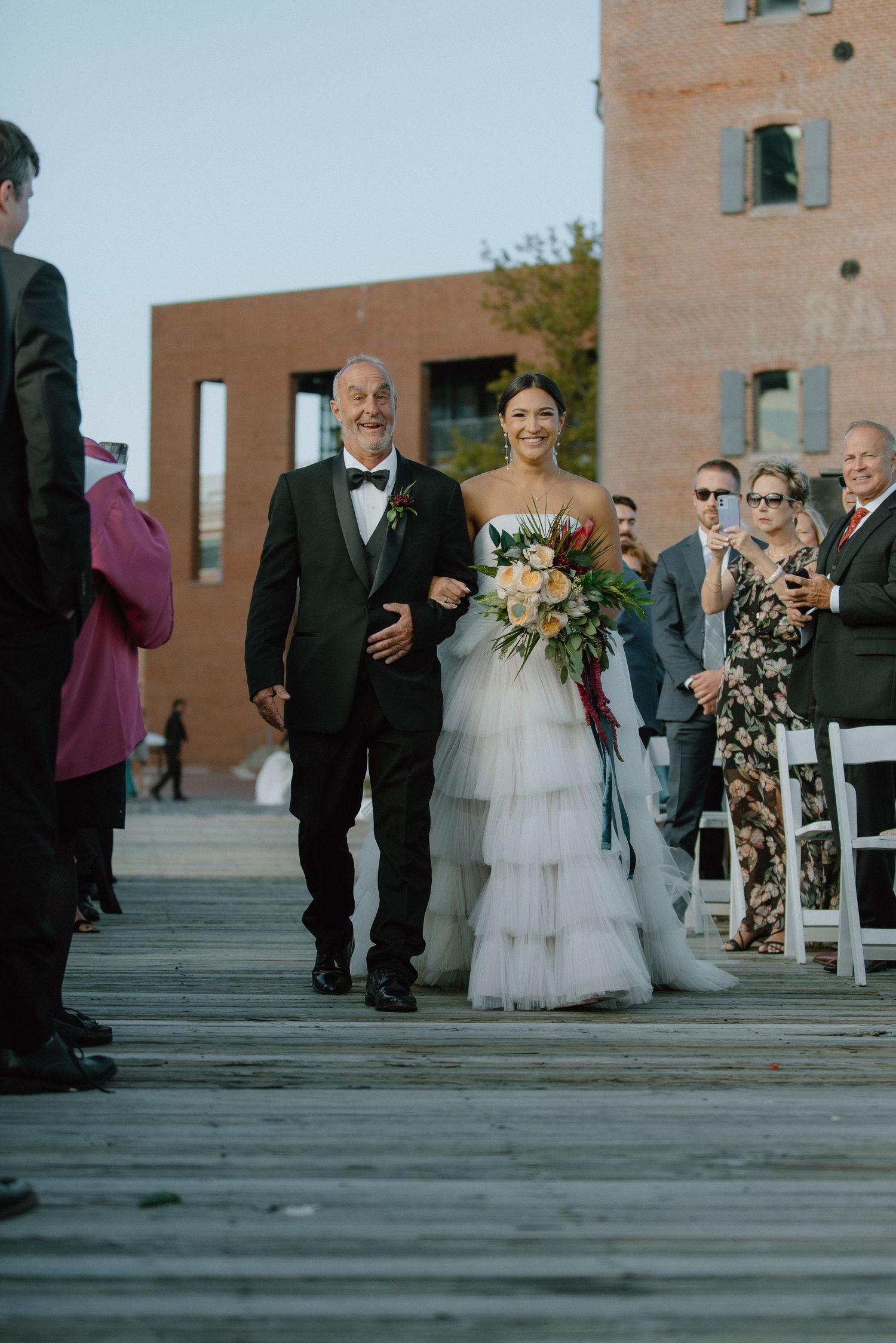 A wedding ceremony taking place on a wooden boardwalk with guests seated and a stately brick building in the background.