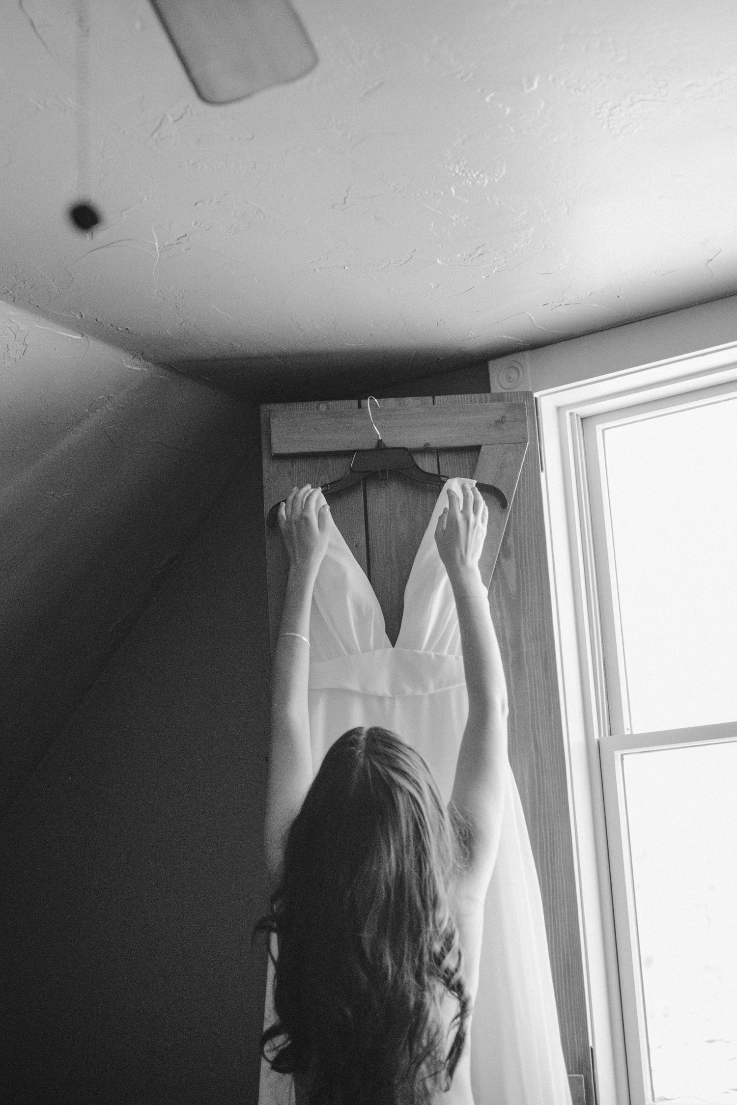 Elegant white wedding dress hanging by window as someone reaches up to adjust it in black and white photo.