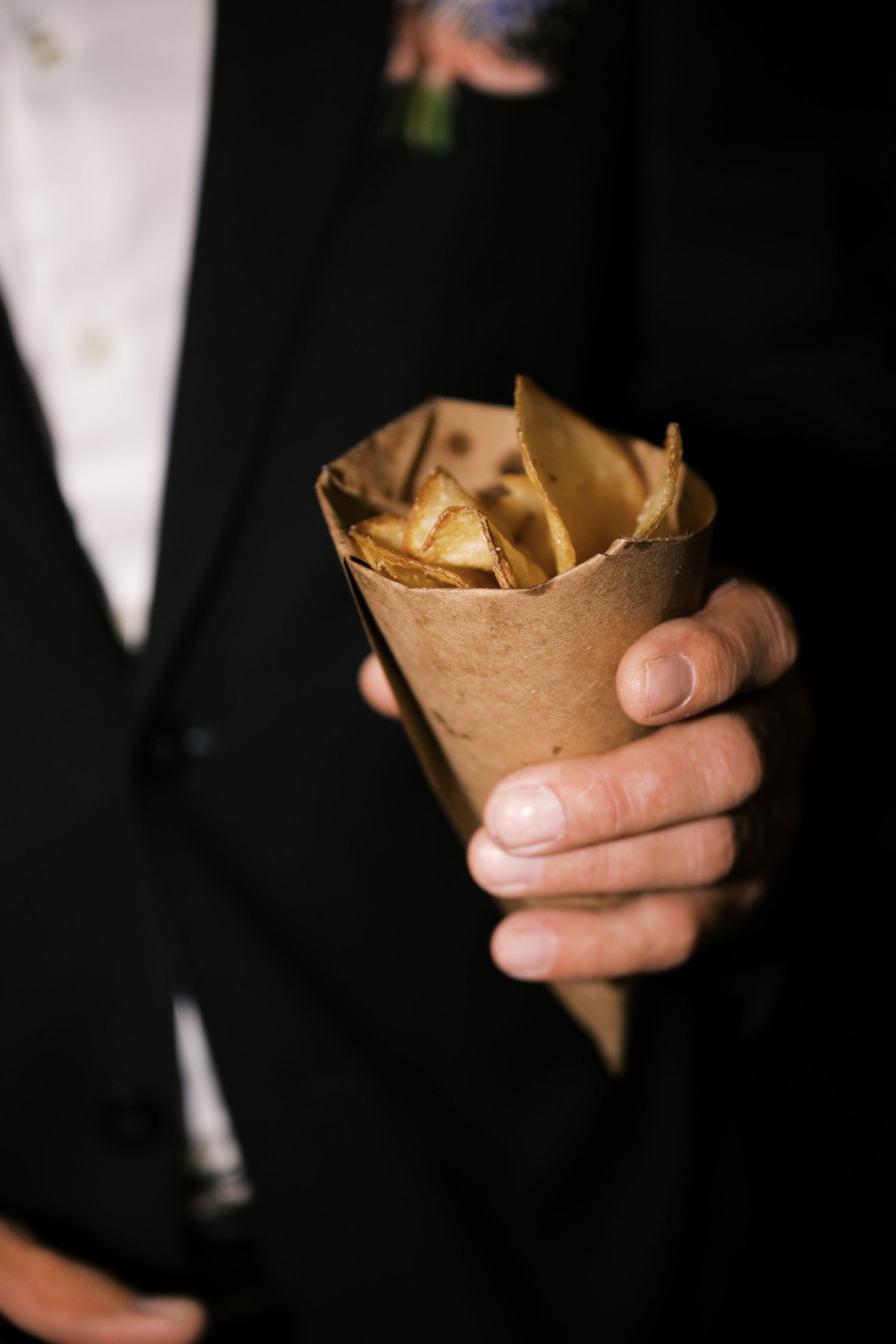 Hand holding wrapped appetizers in brown paper cone at formal event.