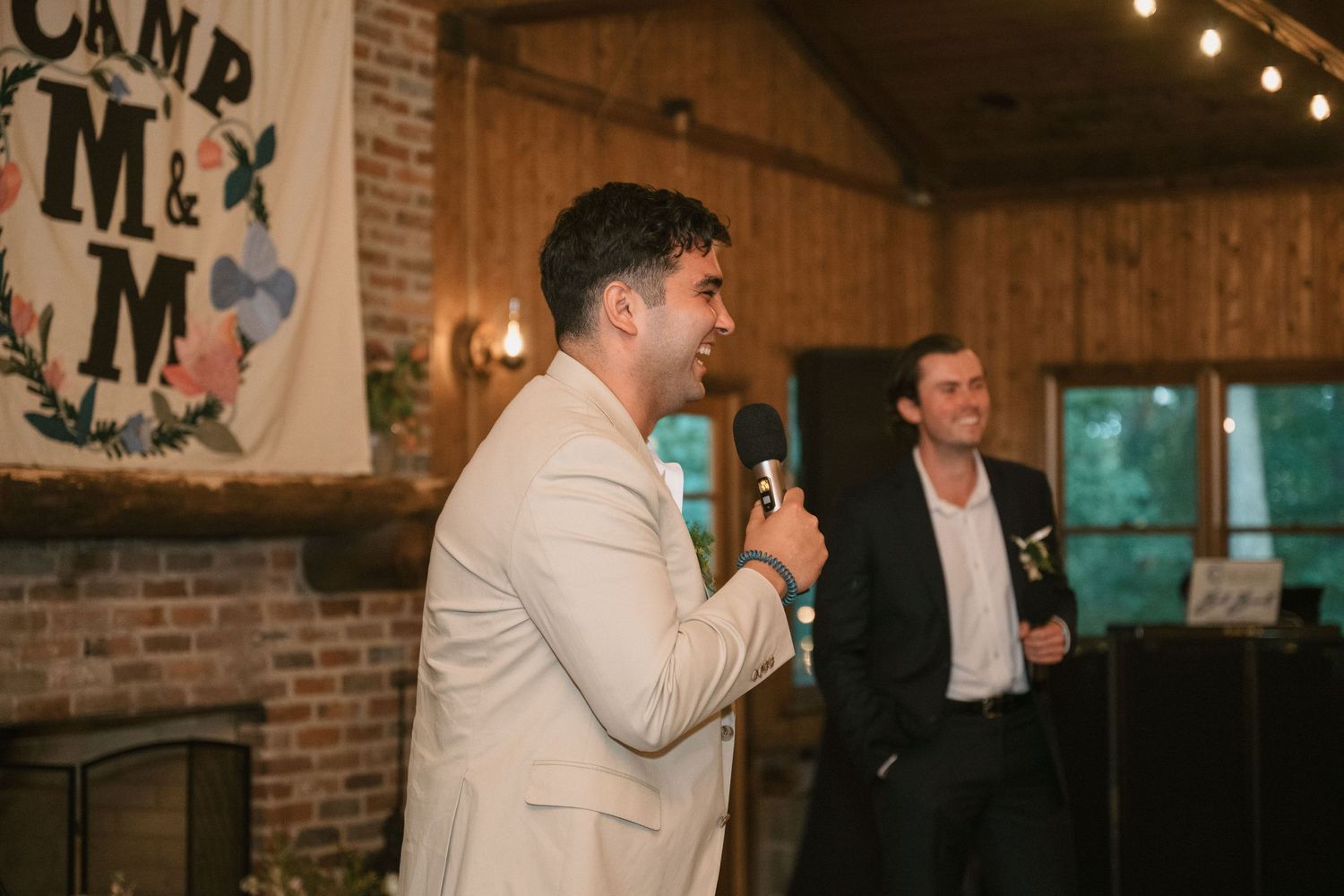 Guest giving speech at rustic venue with Camp M banner in background.