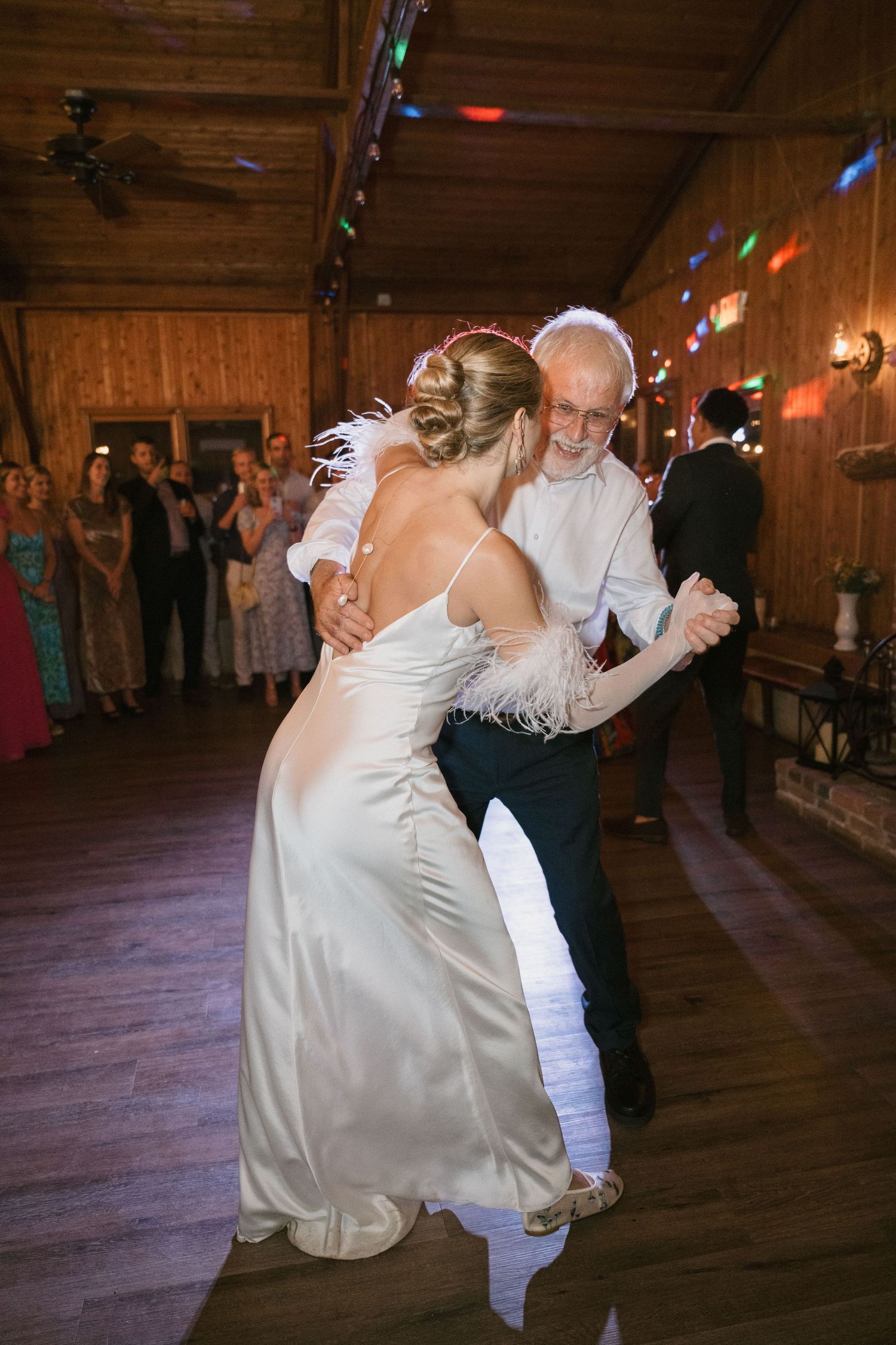 A bride wearing feather details dances with an older gentleman at a rustic venue.