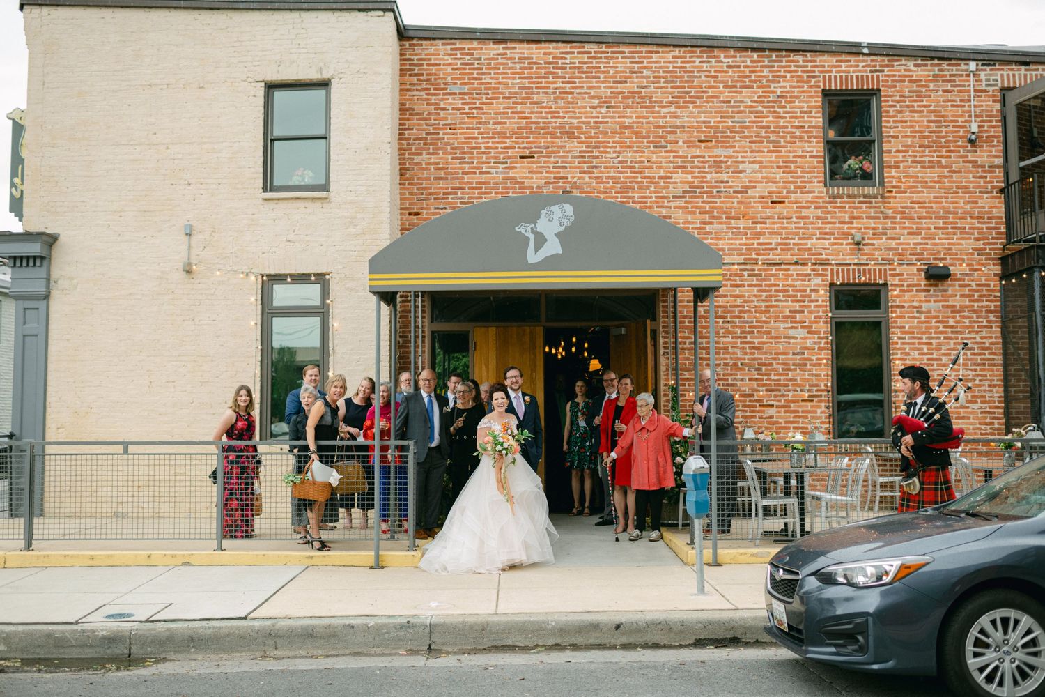 A wedding celebration spills onto the sidewalk outside a rustic brick building entrance.