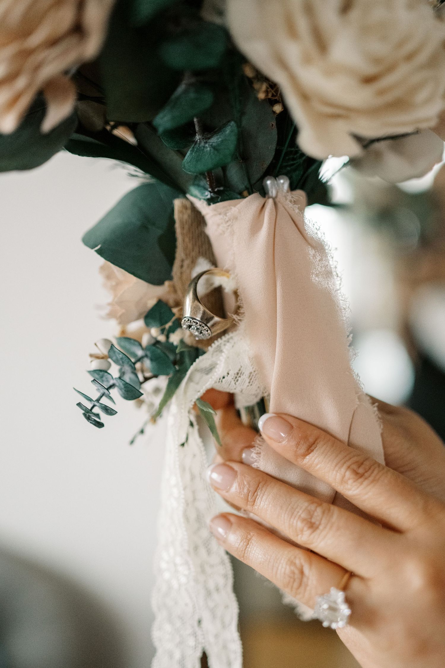 Close-up view of hands holding a delicate bridal bouquet with white roses and green eucalyptus wrapped in lace.