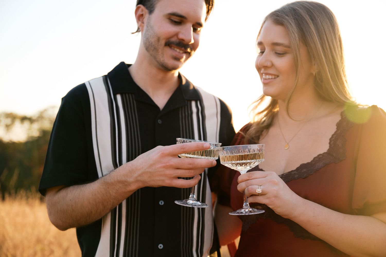 A couple toasts with champagne glasses while enjoying a romantic moment in a sunlit field at dusk.