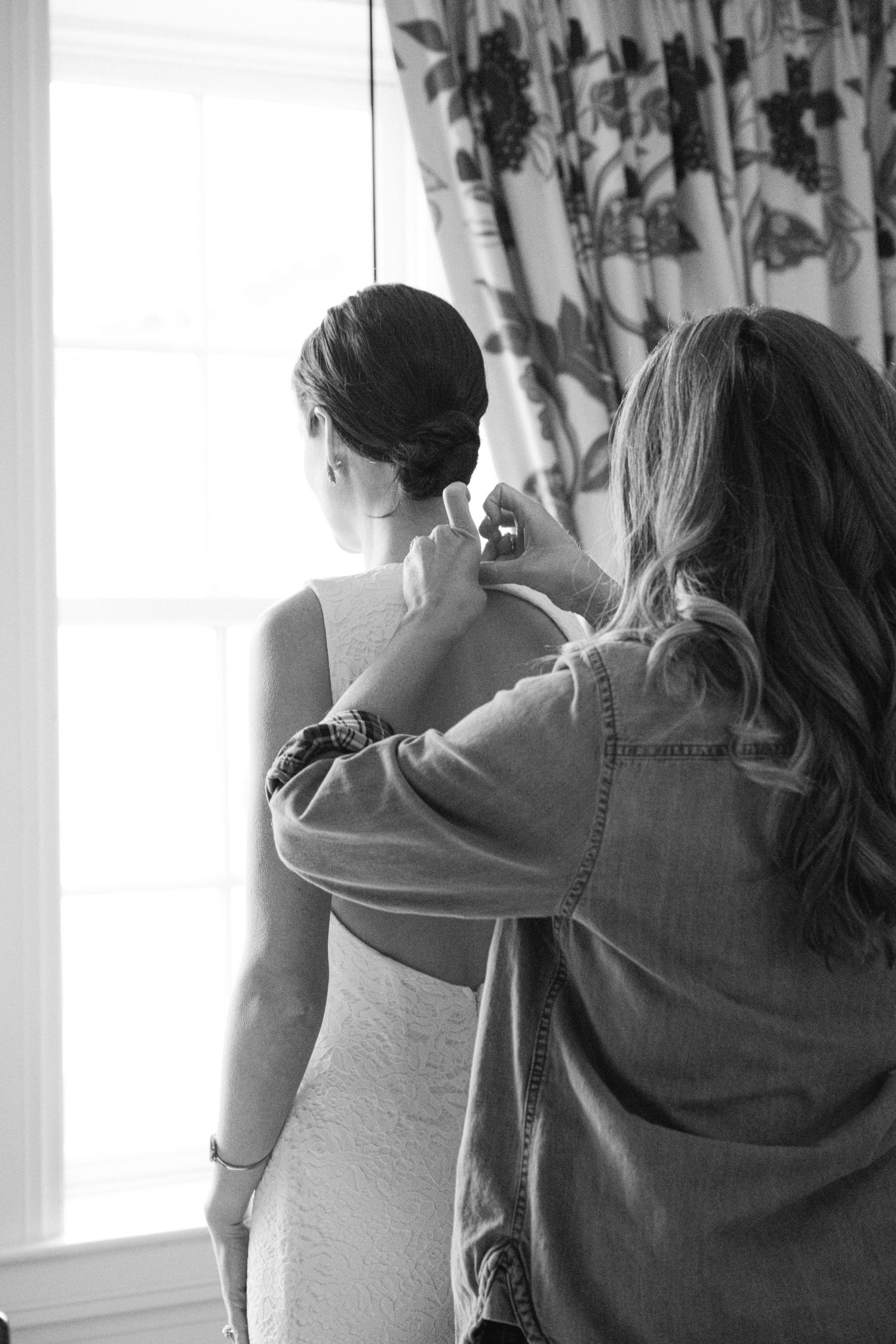 Getting ready scene as a white dress is adjusted near a window with floral curtains.