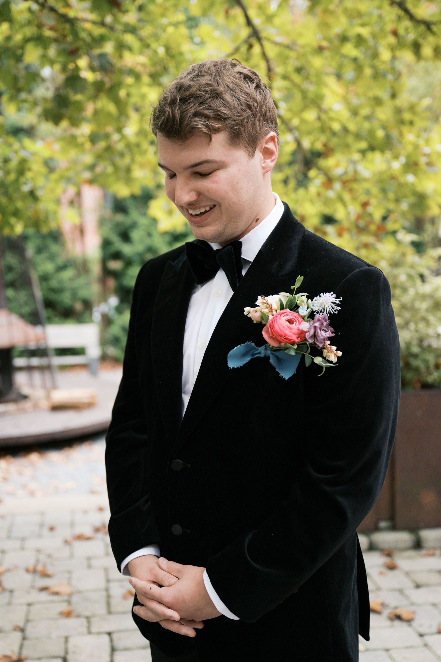 A groom in a black velvet tuxedo with a pink and white floral boutonniere smiles while looking down.