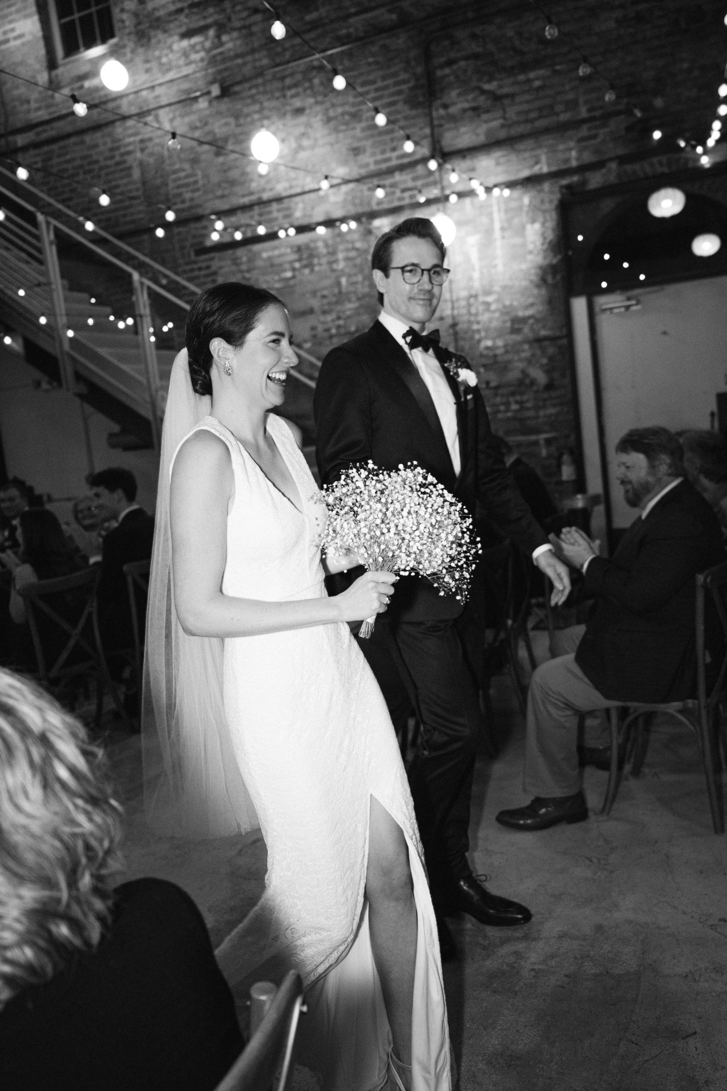 Black and white wedding photo showing a couple in formal attire with string lights in an industrial venue space.