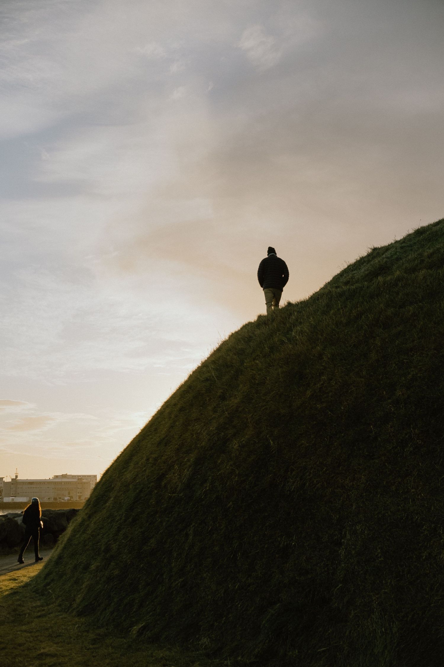 Silhouetted person standing atop steep grassy hill during golden hour.