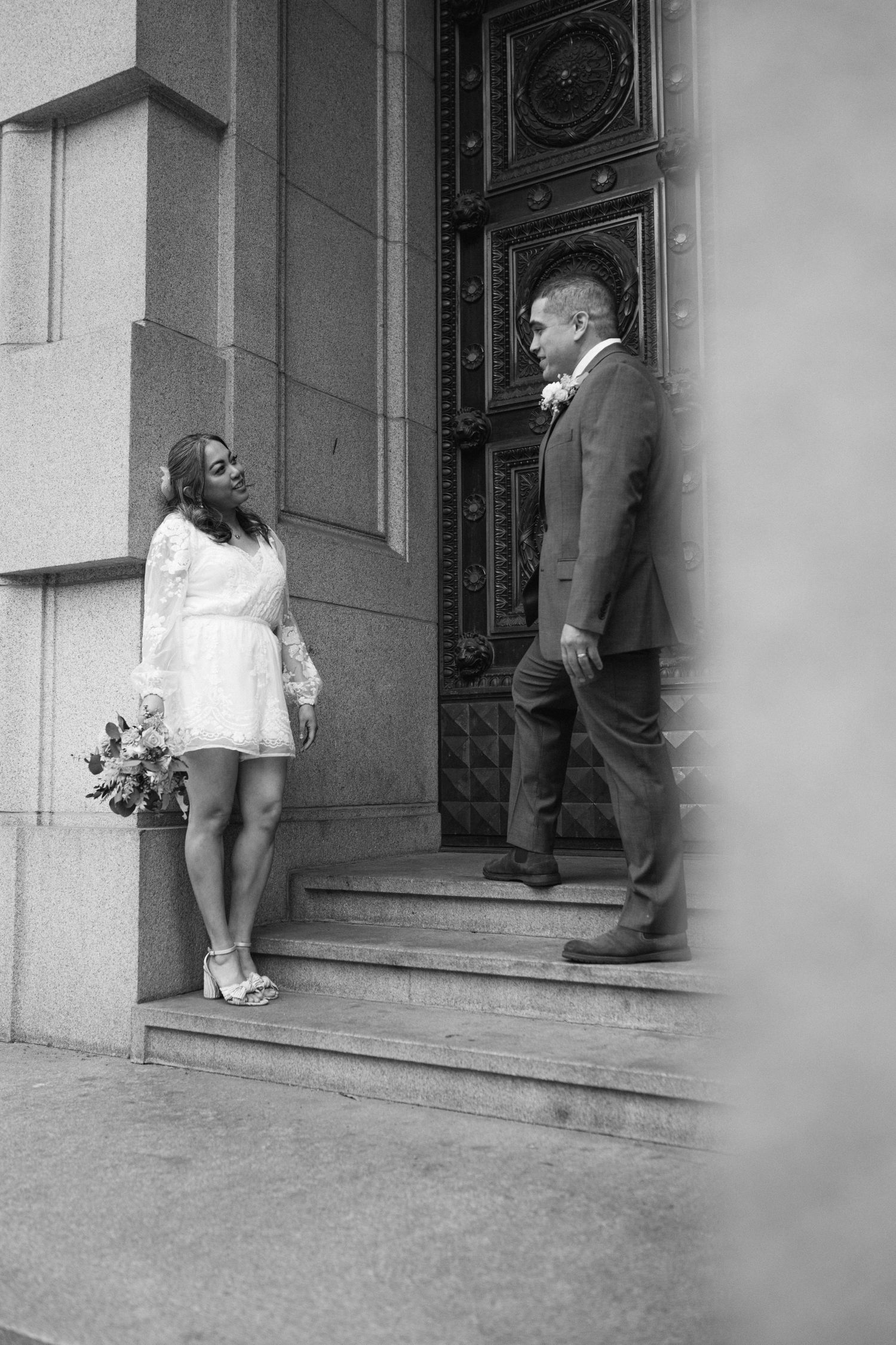 A couple stands on stone steps by an ornate door in black and white, with the woman holding a bouquet in a white dress.