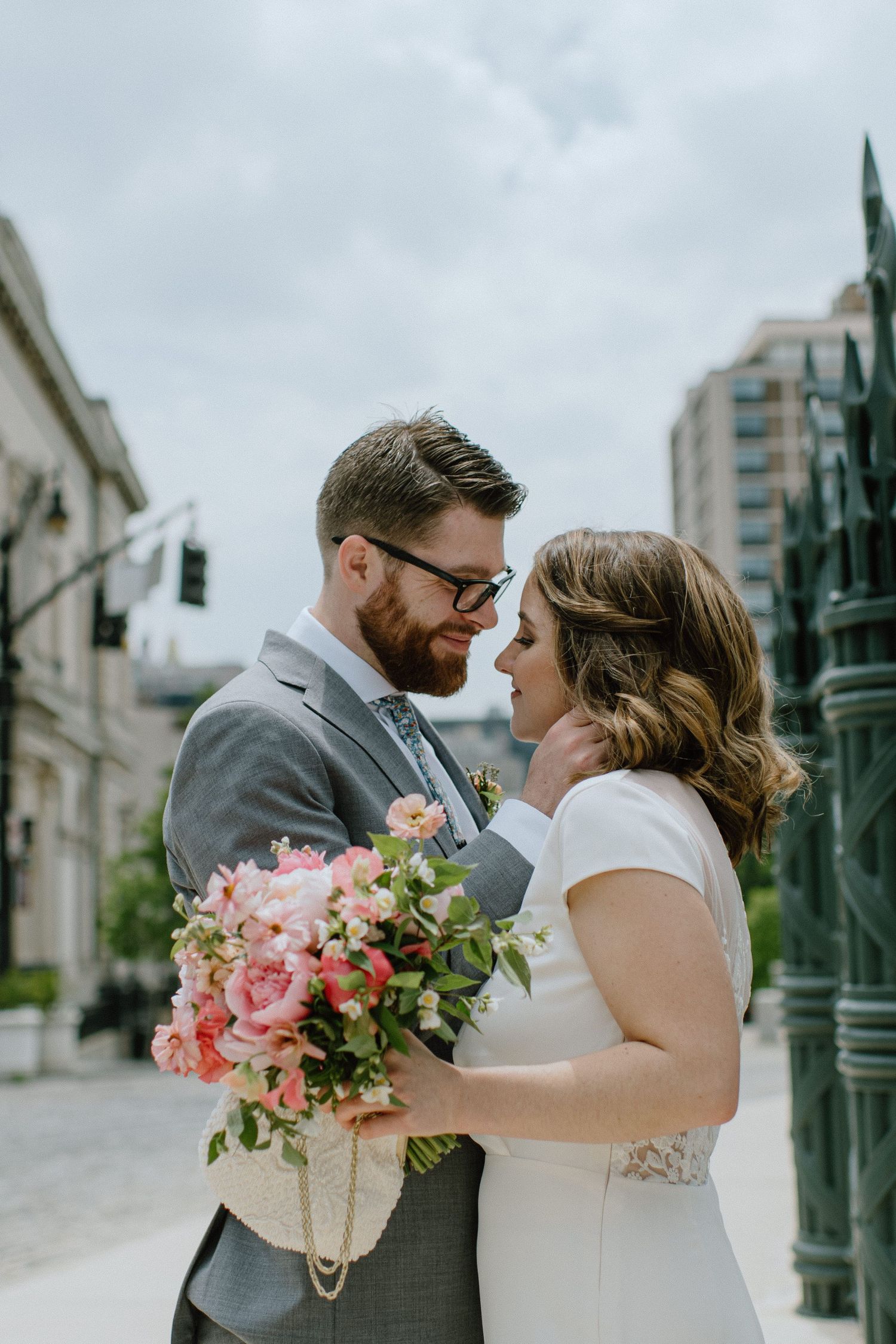 Intimate moment between newlyweds holding pink bouquet against urban backdrop.
