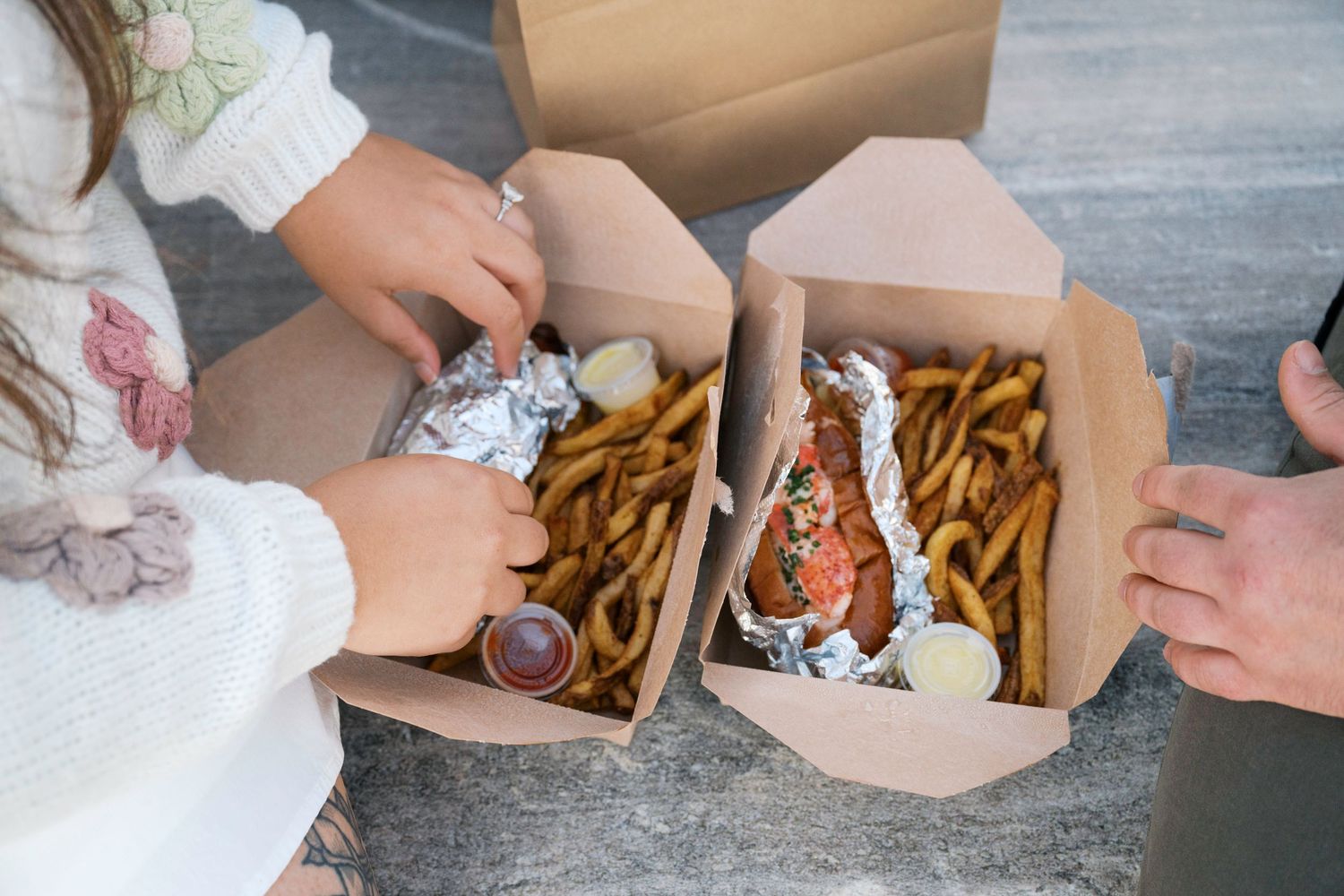 Hands reaching for takeout containers filled with french fries and dipping sauce on a concrete surface.
