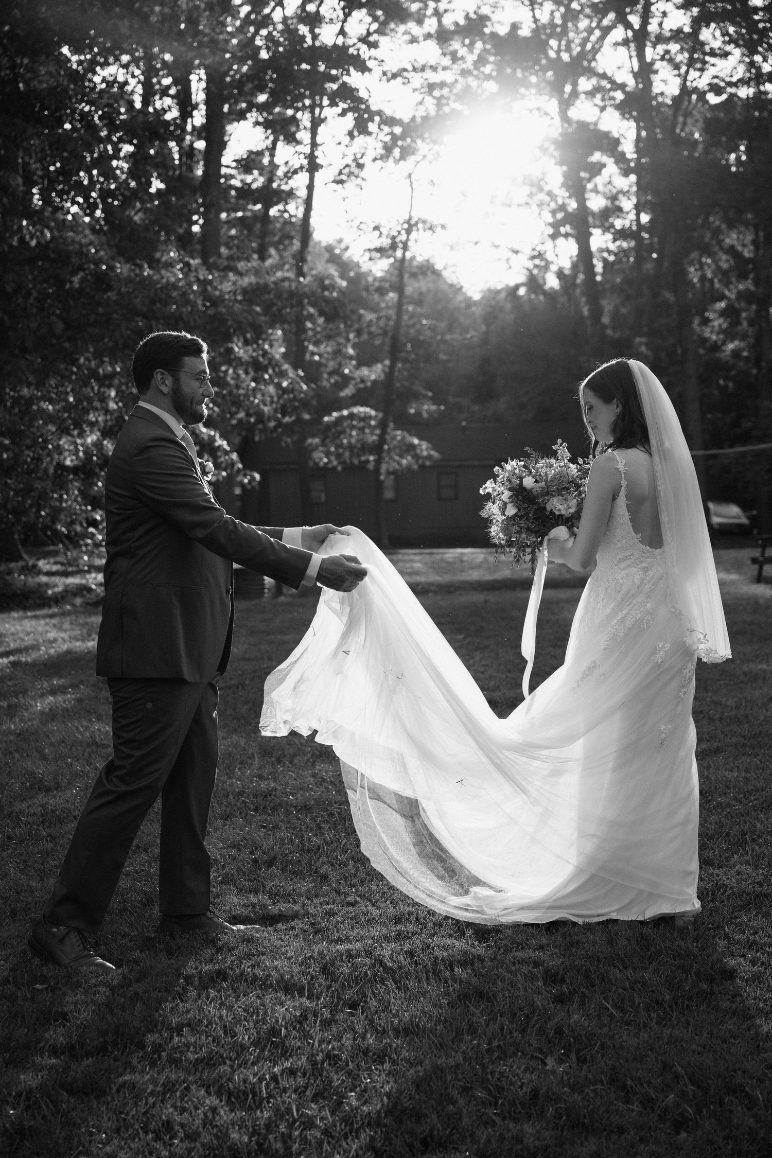 A romantic black and white wedding photo showing a couple standing in a sunlit forest with the bride's dress flowing in the breeze.