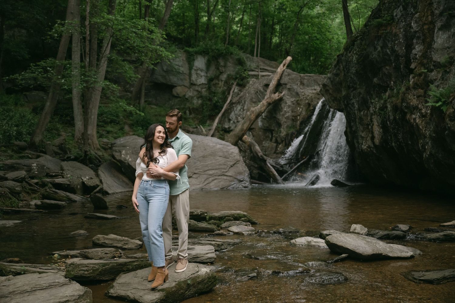 A couple enjoys a tender moment along a rocky stream with a waterfall in the background.