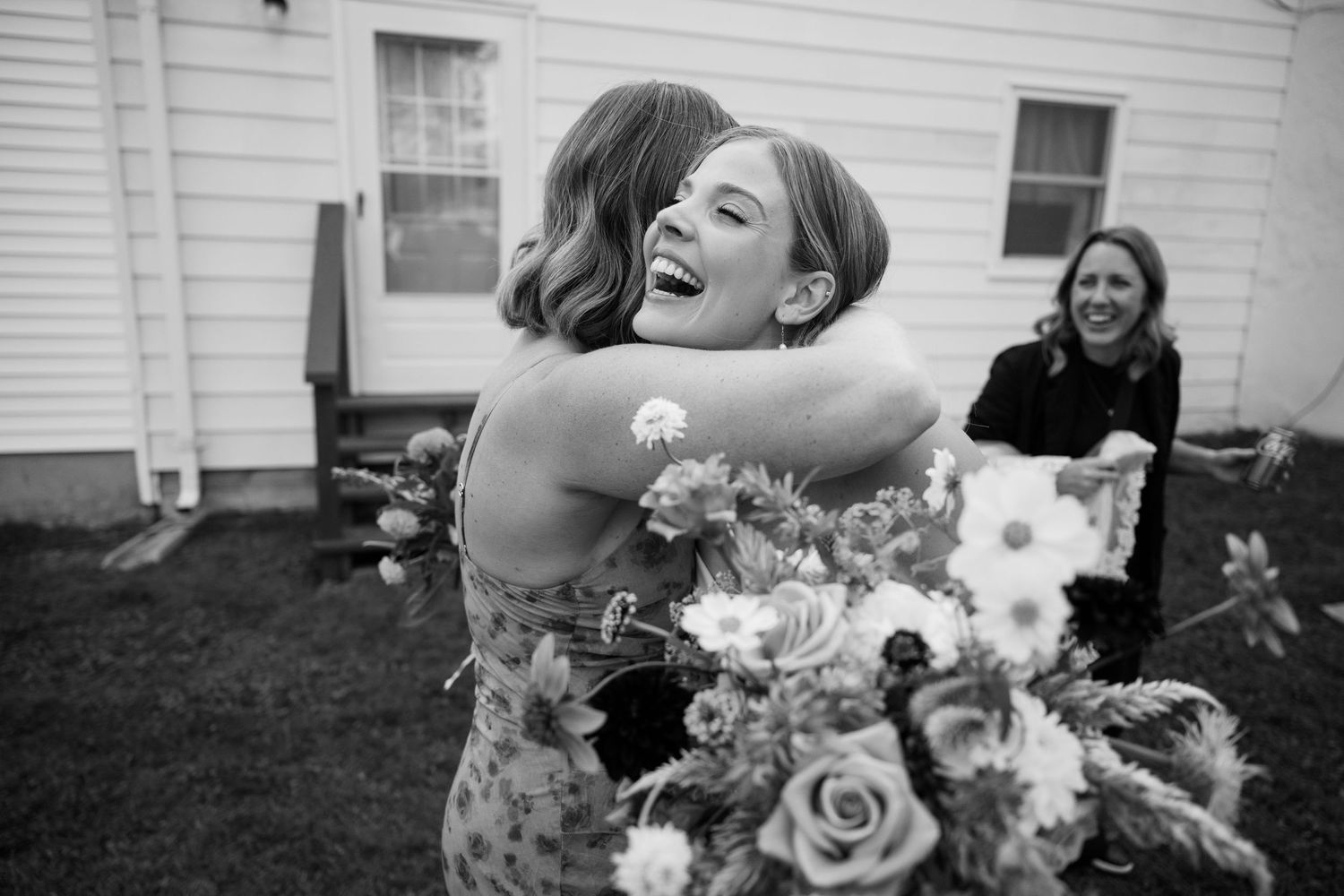 Two women share a joyful hug at a wedding, one holding a floral bouquet, with a smiling guest in the background.