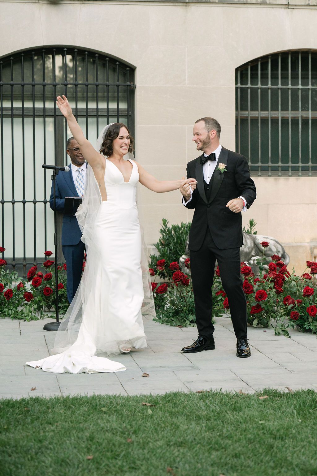A joyful wedding couple celebrates on a lawn with red flowers and arched windows in the background.