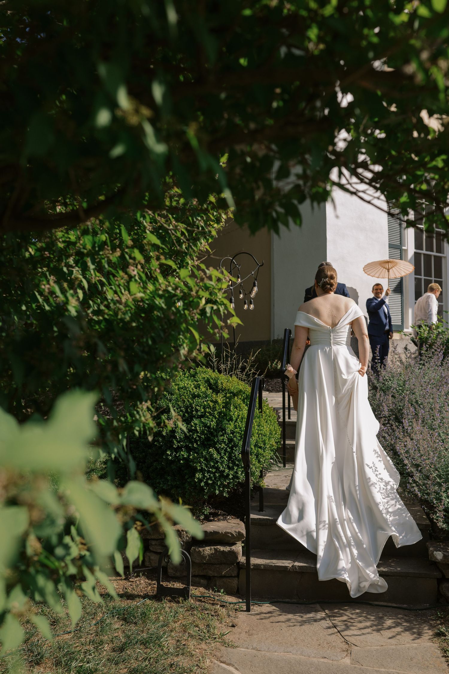 A bride in a white gown walks along a garden path surrounded by lush greenery.