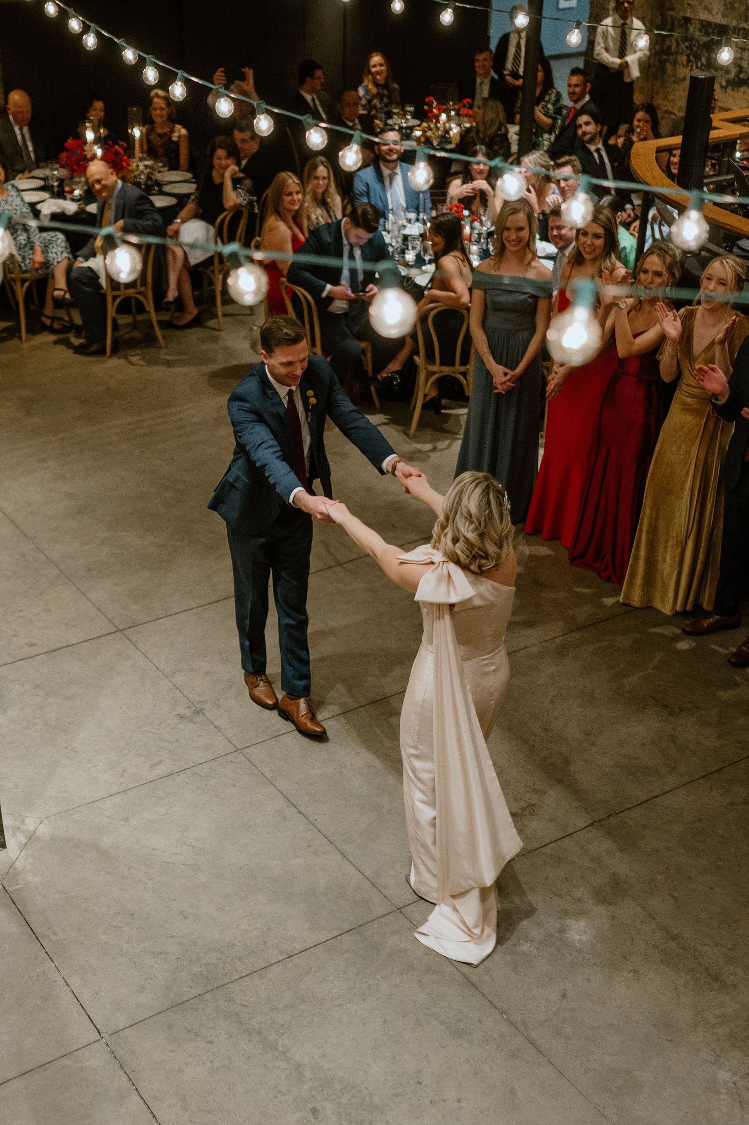 Dancing couple surrounded by guests in formal attire under string lights at reception.
