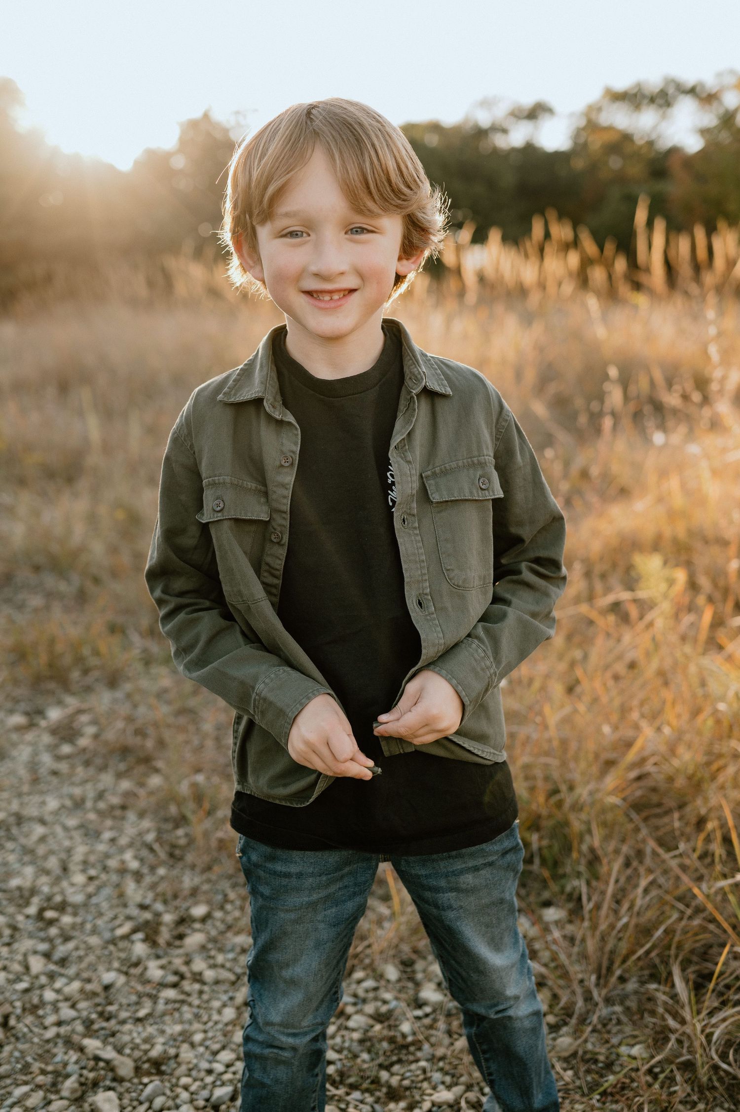 A child stands outdoors wearing a green jacket and black shirt at golden hour with tall grass in the background.