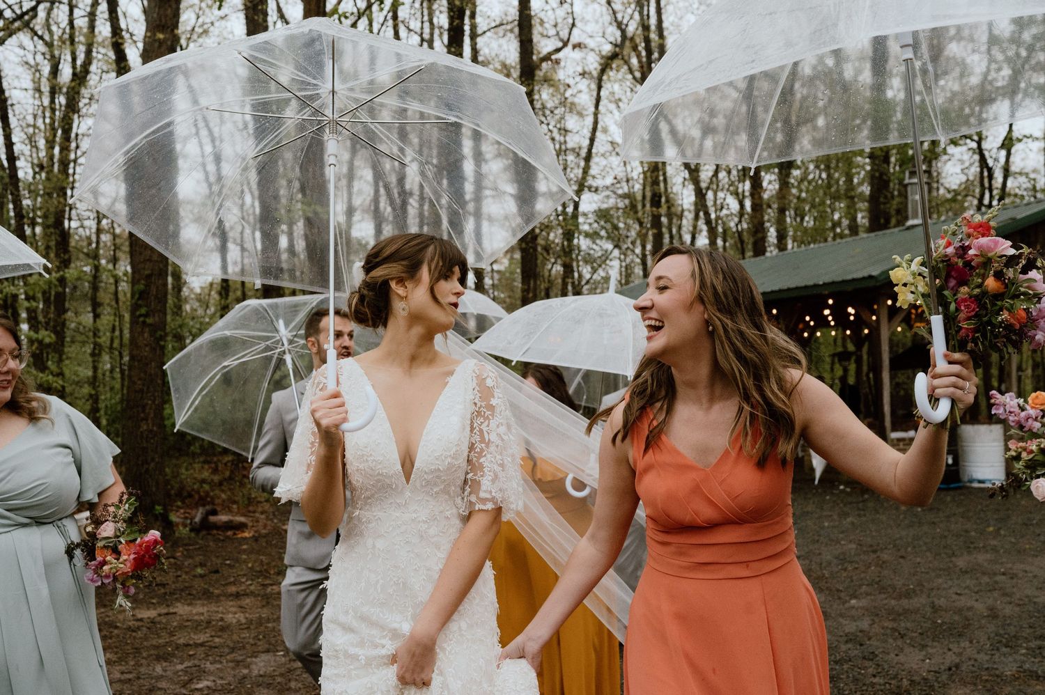 A bride and bridesmaid share a laugh while holding clear umbrellas during a rainy wedding celebration.