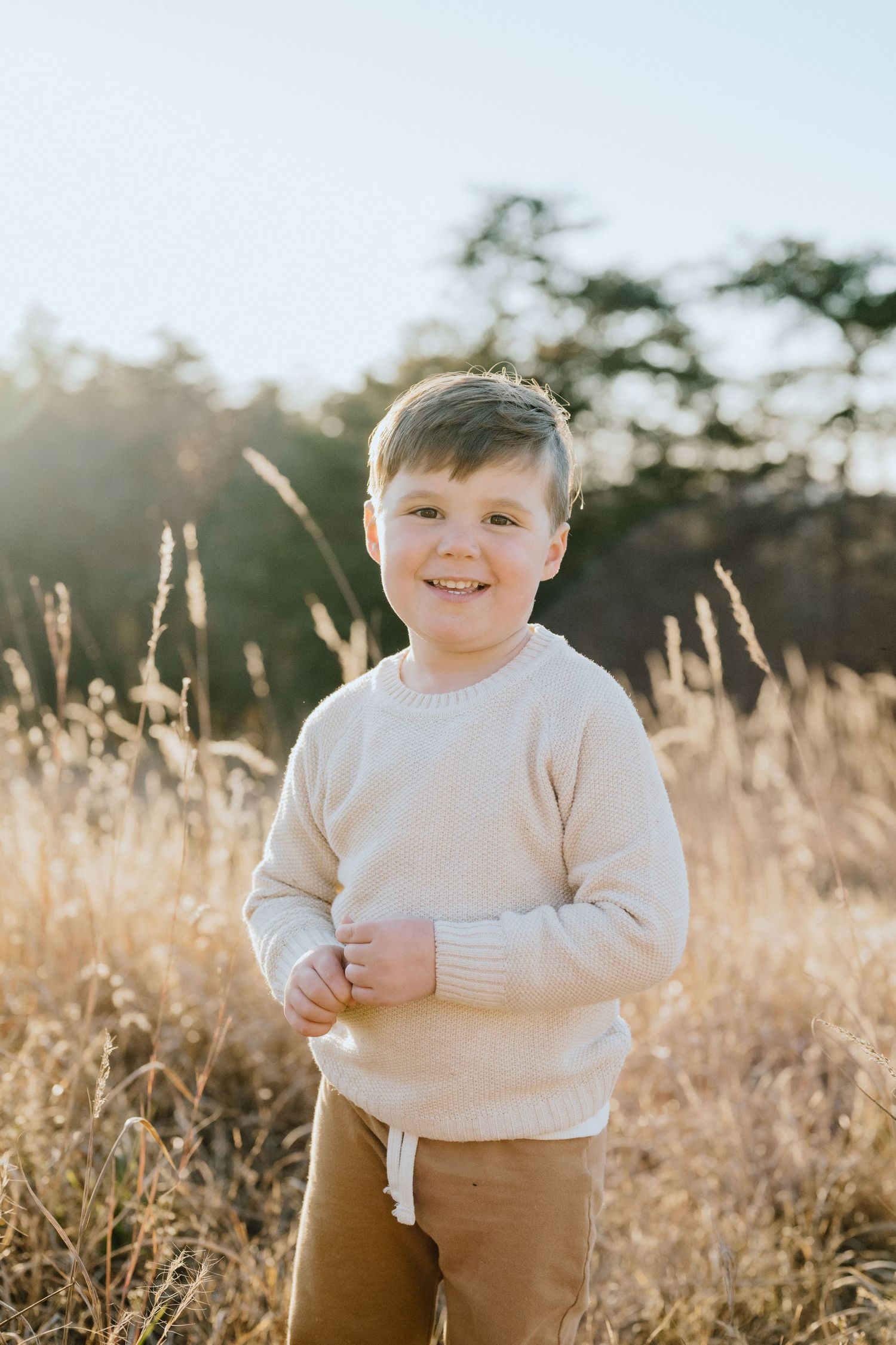 Child in cream sweater smiling while standing in sunlit field of tall grass.