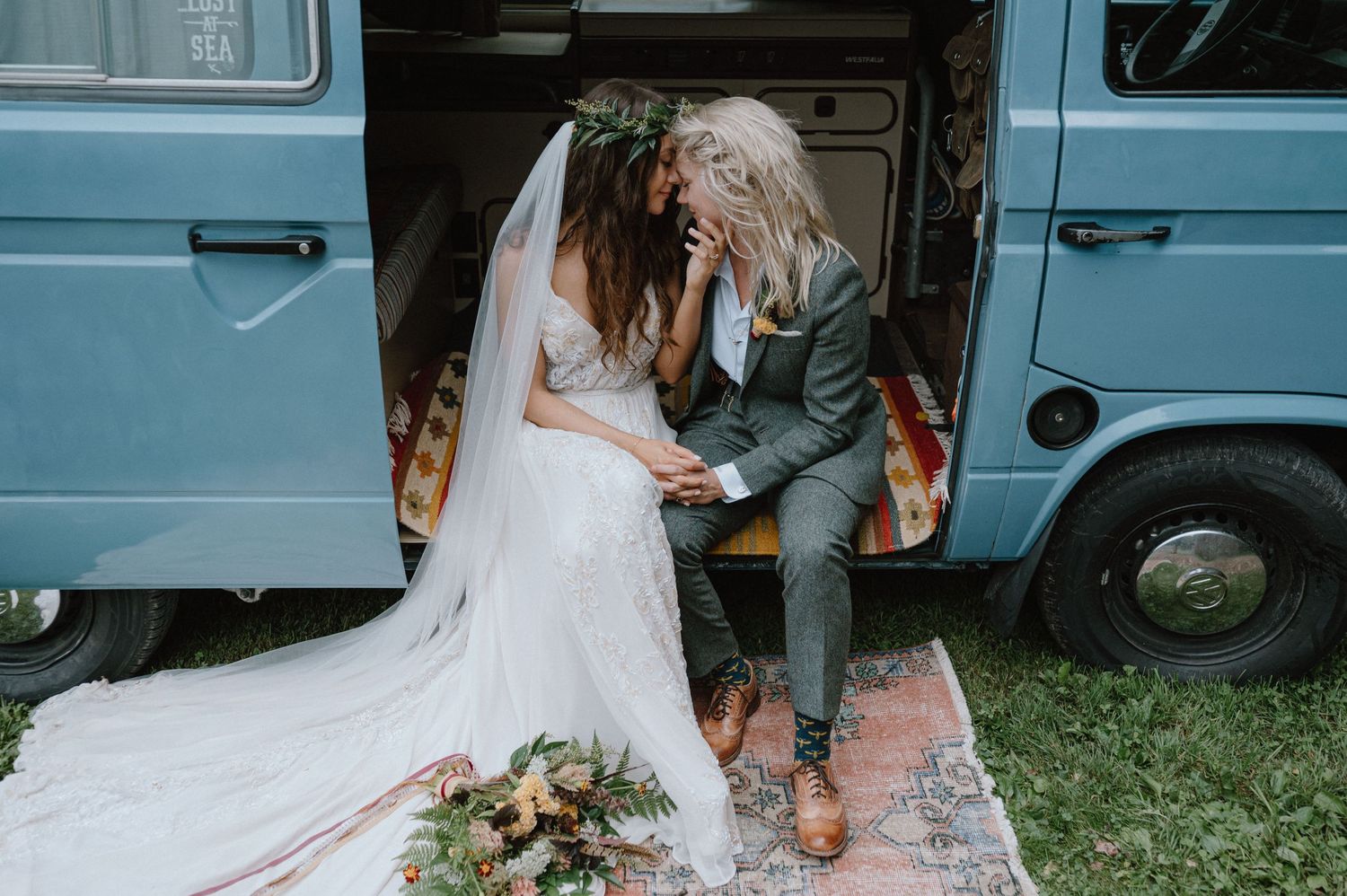 A couple sits in the doorway of a blue vintage VW van in their wedding attire.