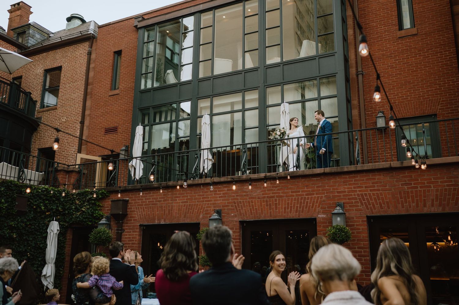 Wedding guests gather below a modern glass balcony on a brick building.