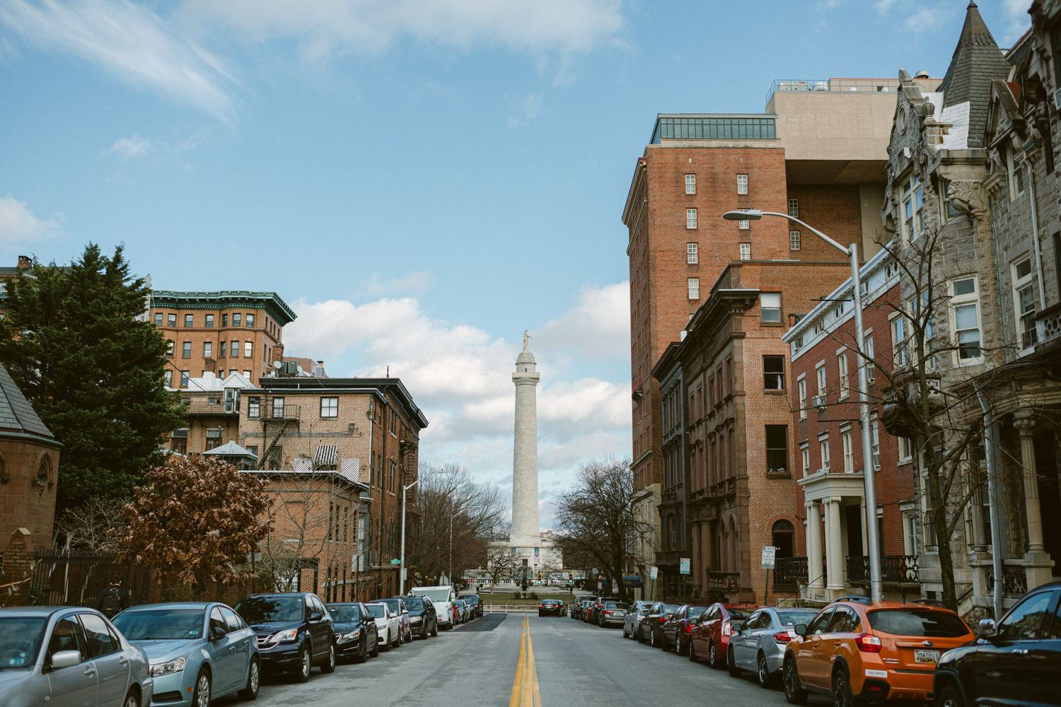 Historic street view with Washington Monument visible between brick buildings and parked cars.