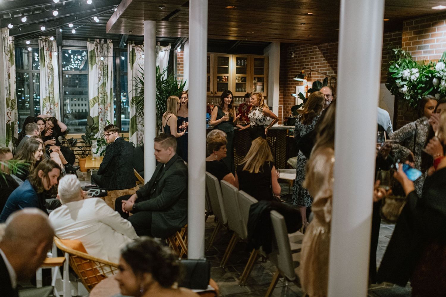 Crowded indoor reception venue with guests seated at tables under string lights.