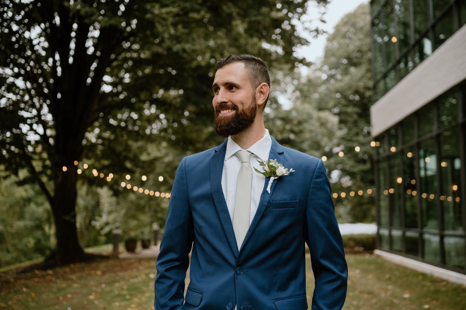 Groom in navy blue suit with boutonniere smiling outdoors with string lights and greenery in background.