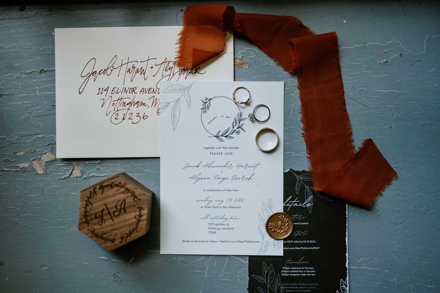 Wedding invitation with burgundy ribbon, vintage wax seal, and gold wedding rings on blue surface.