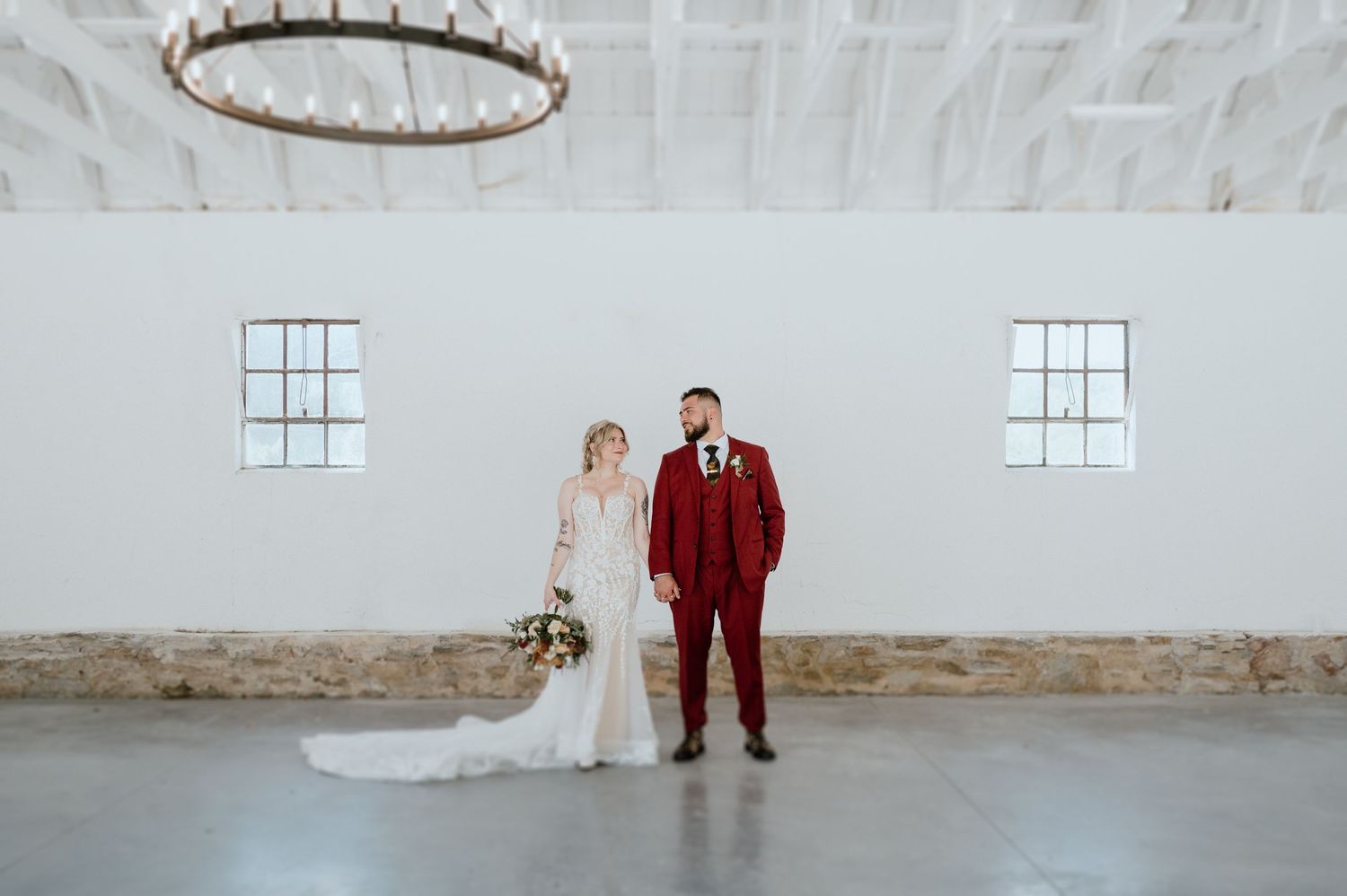 A wedding couple poses in a minimalist white room with stone walls and a circular chandelier overhead.