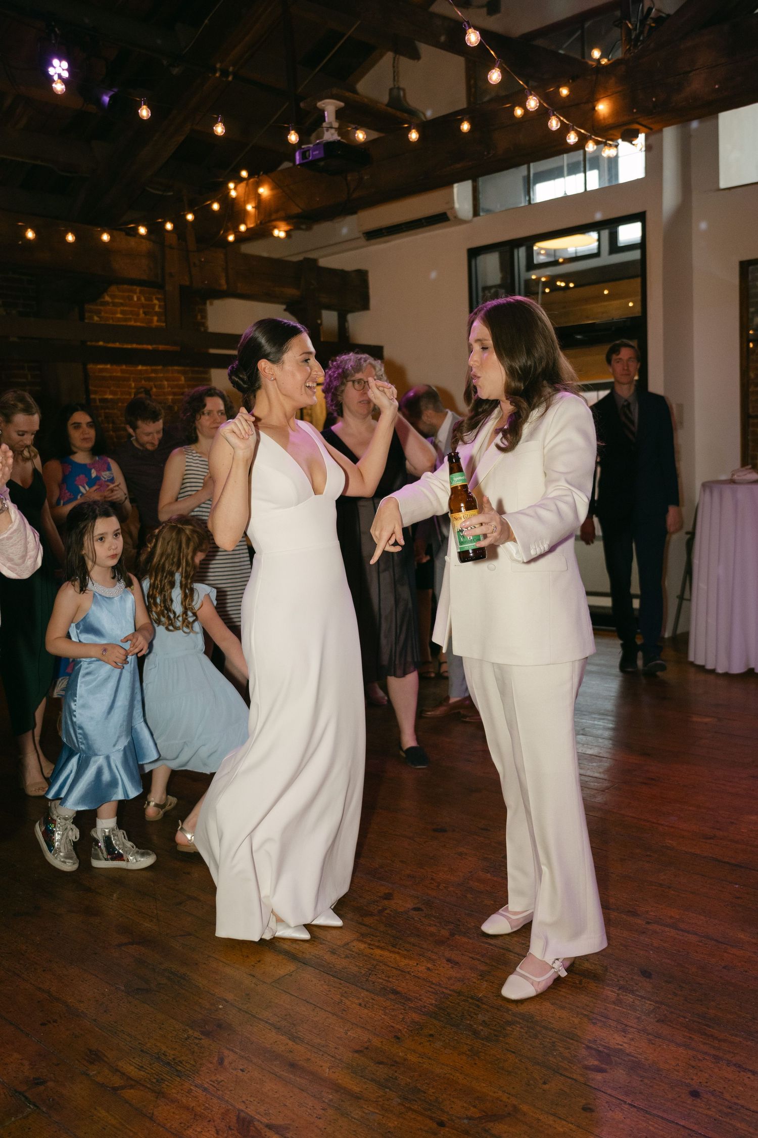 Wedding guests dance together on a rustic wooden dance floor under glowing string lights at an indoor reception venue.
