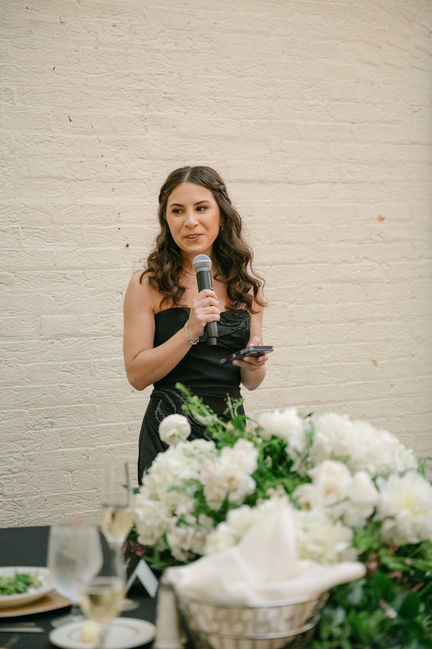 Someone in a black dress gives a speech at an elegant reception with white floral centerpieces.