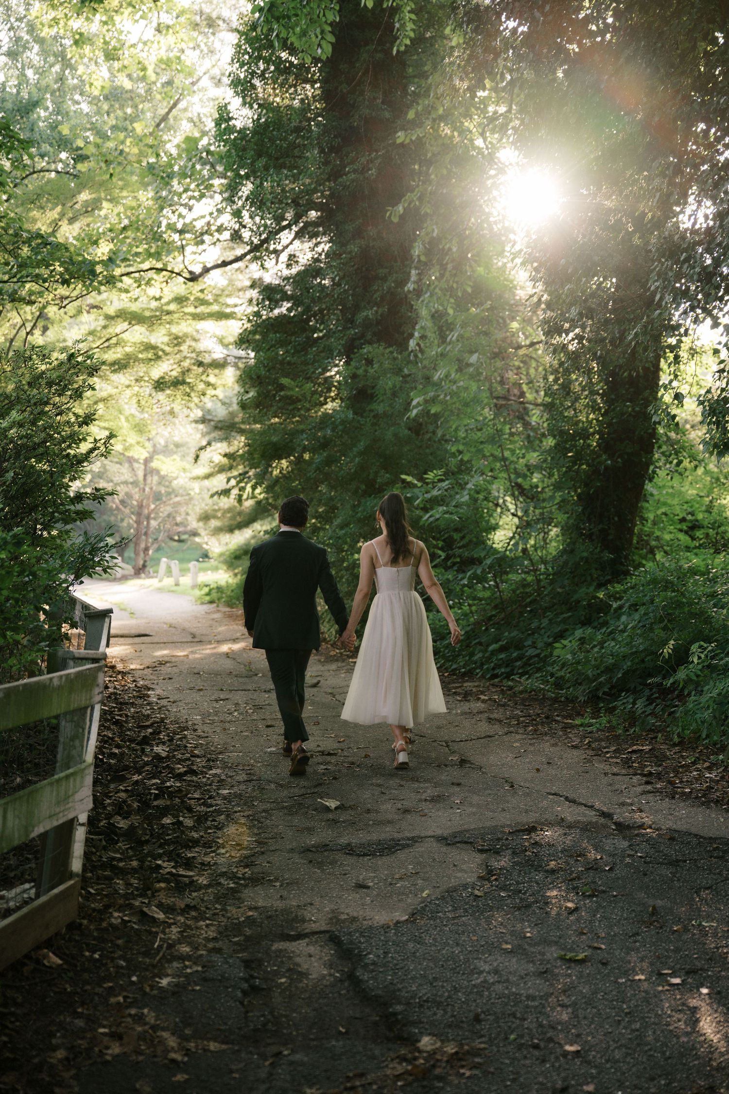 A couple walks hand in hand down a scenic wooded path with sunlight streaming through the trees.