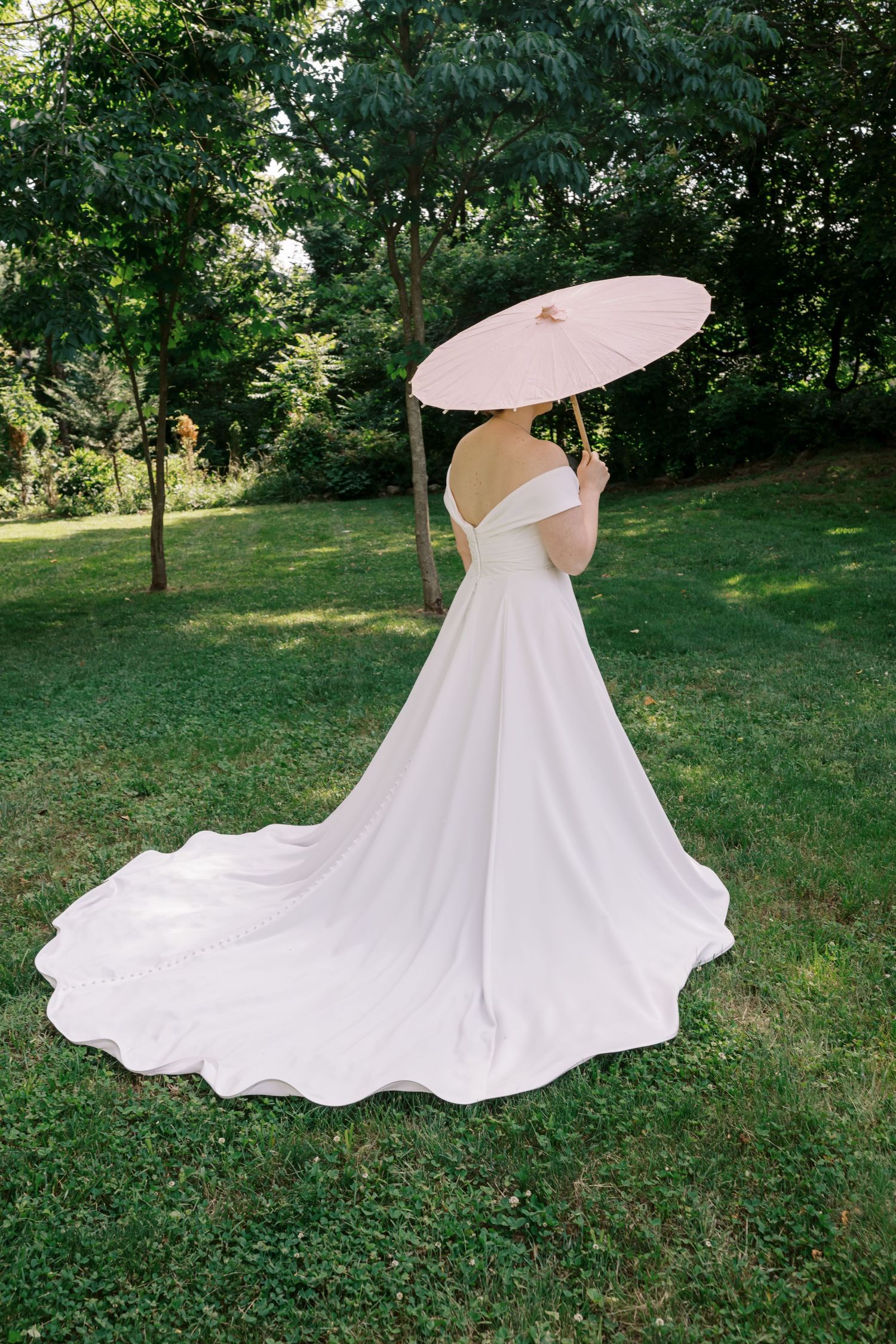 Bride in white wedding dress with long train holding pink parasol in garden setting.