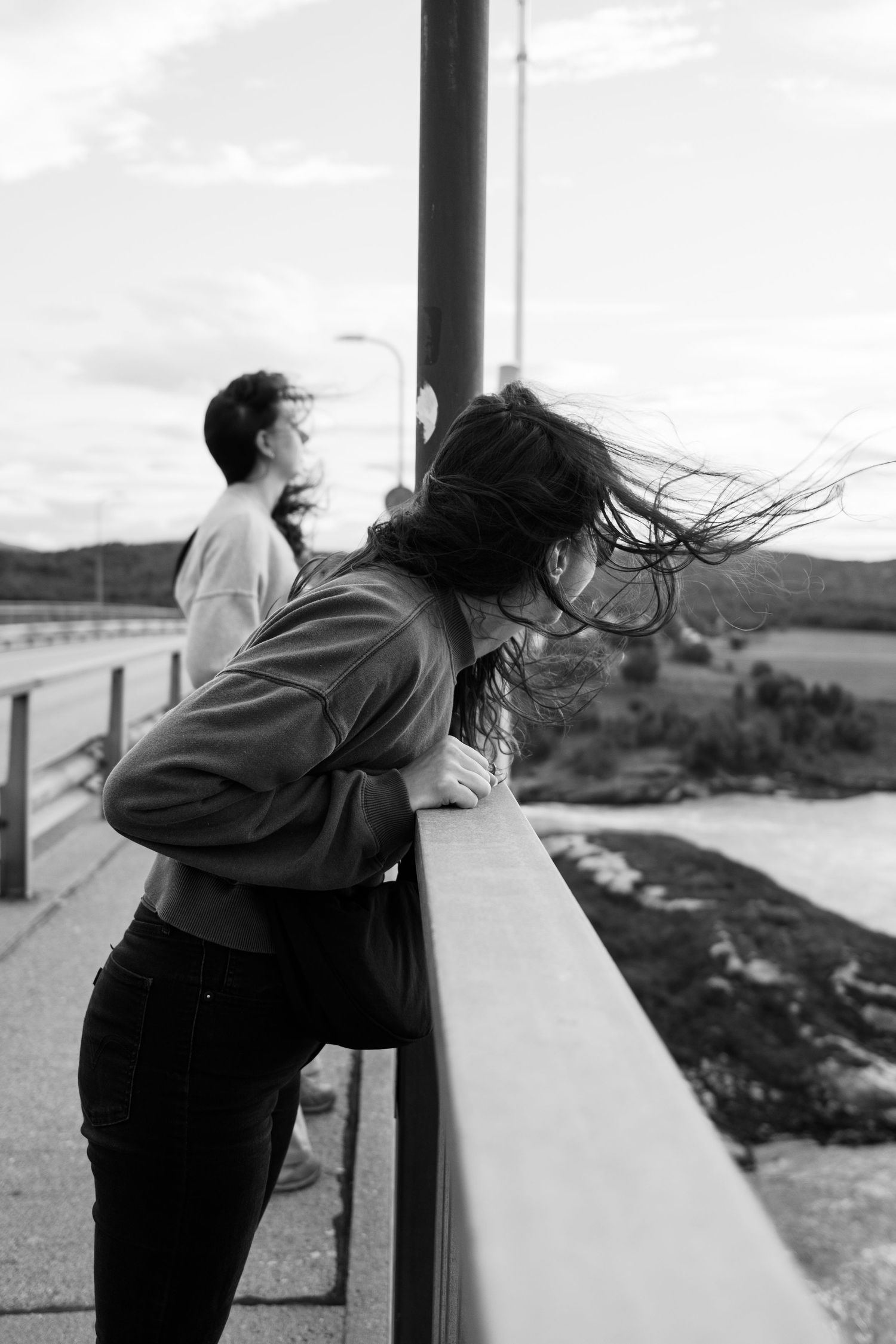 Person leaning over bridge railing with wind-blown hair against scenic coastal backdrop.