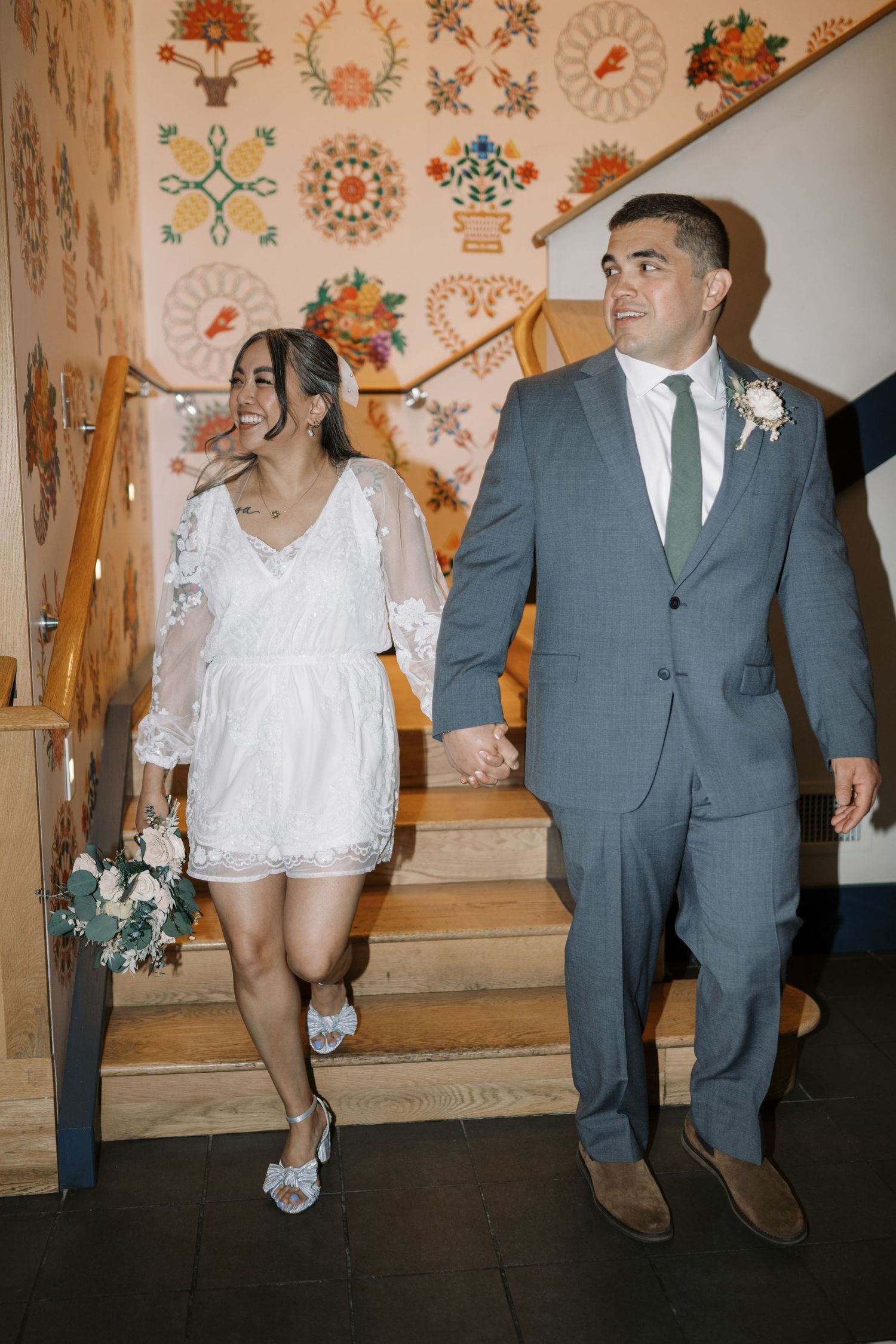 A couple descends stairs with decorative wall art, the bride in a white lace dress and groom in grey suit.