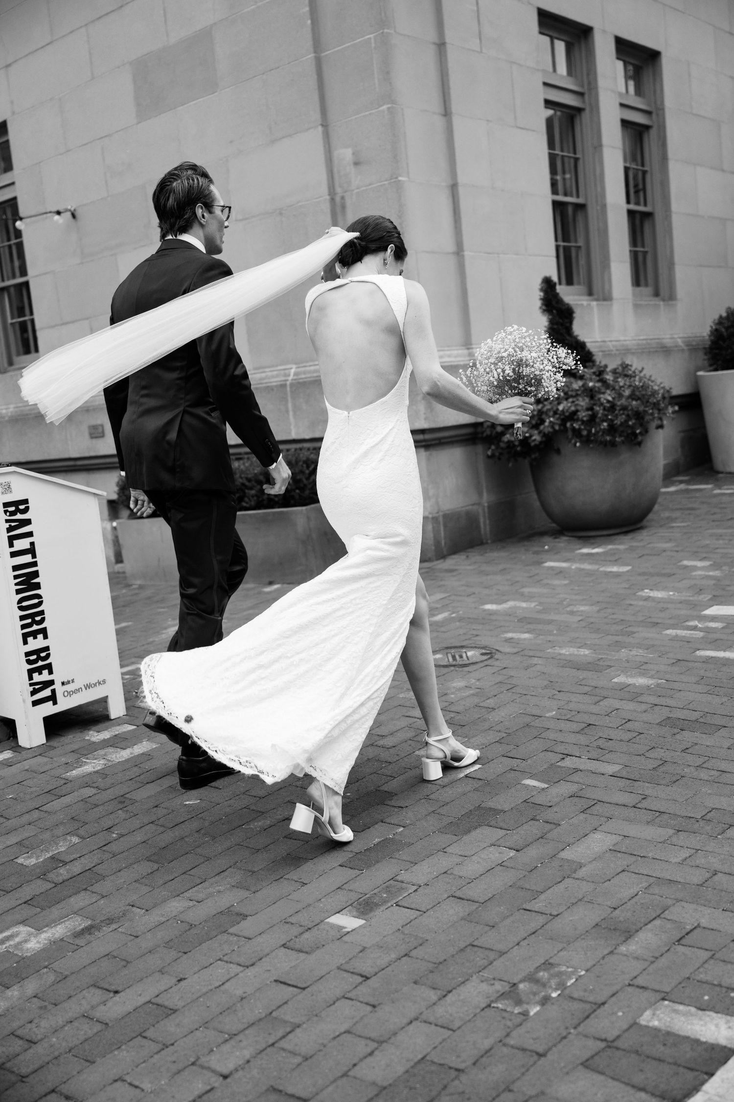 A backless wedding gown flows gracefully as a couple walks together on brick pavement in black and white photography.