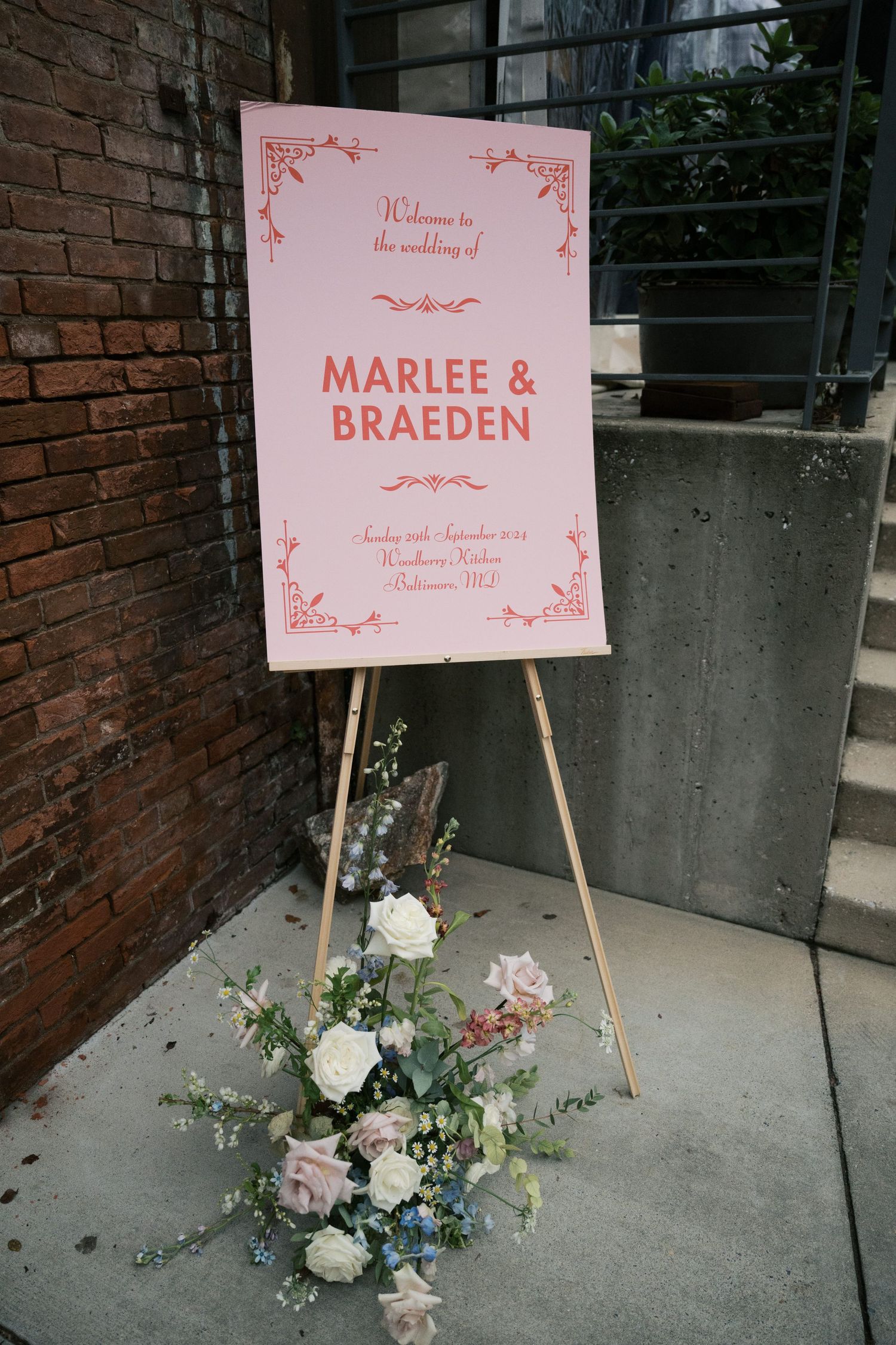 A pink wedding welcome sign for Marlee & Braeden displayed on an easel with floral arrangements.