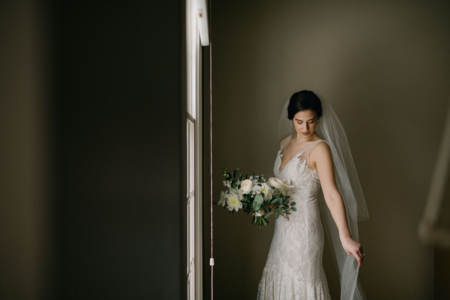 Elegant bridal portrait by window with white bouquet and lace dress.