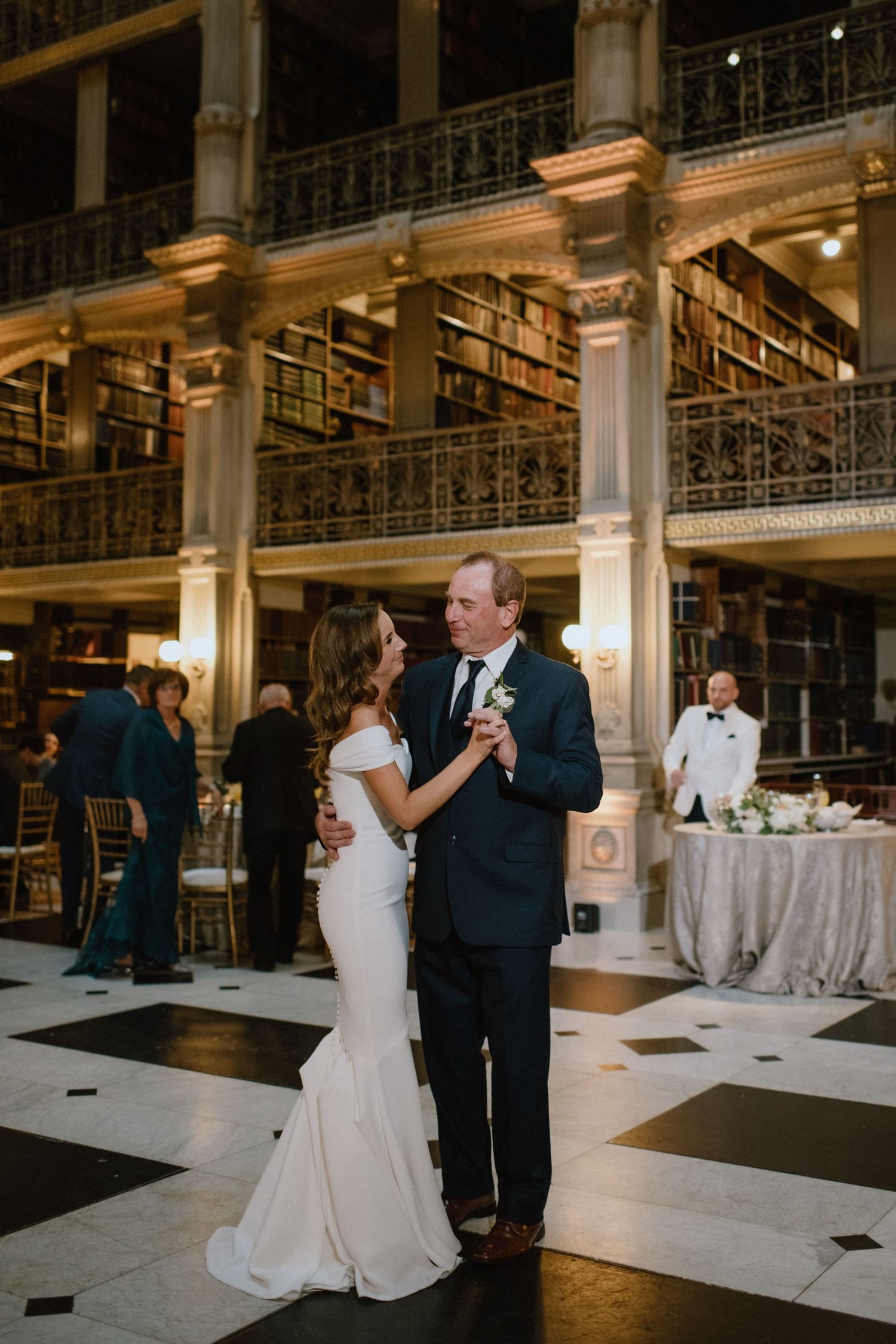 Couple shares first dance on checkered floor of grand historic library with towering bookshelves.