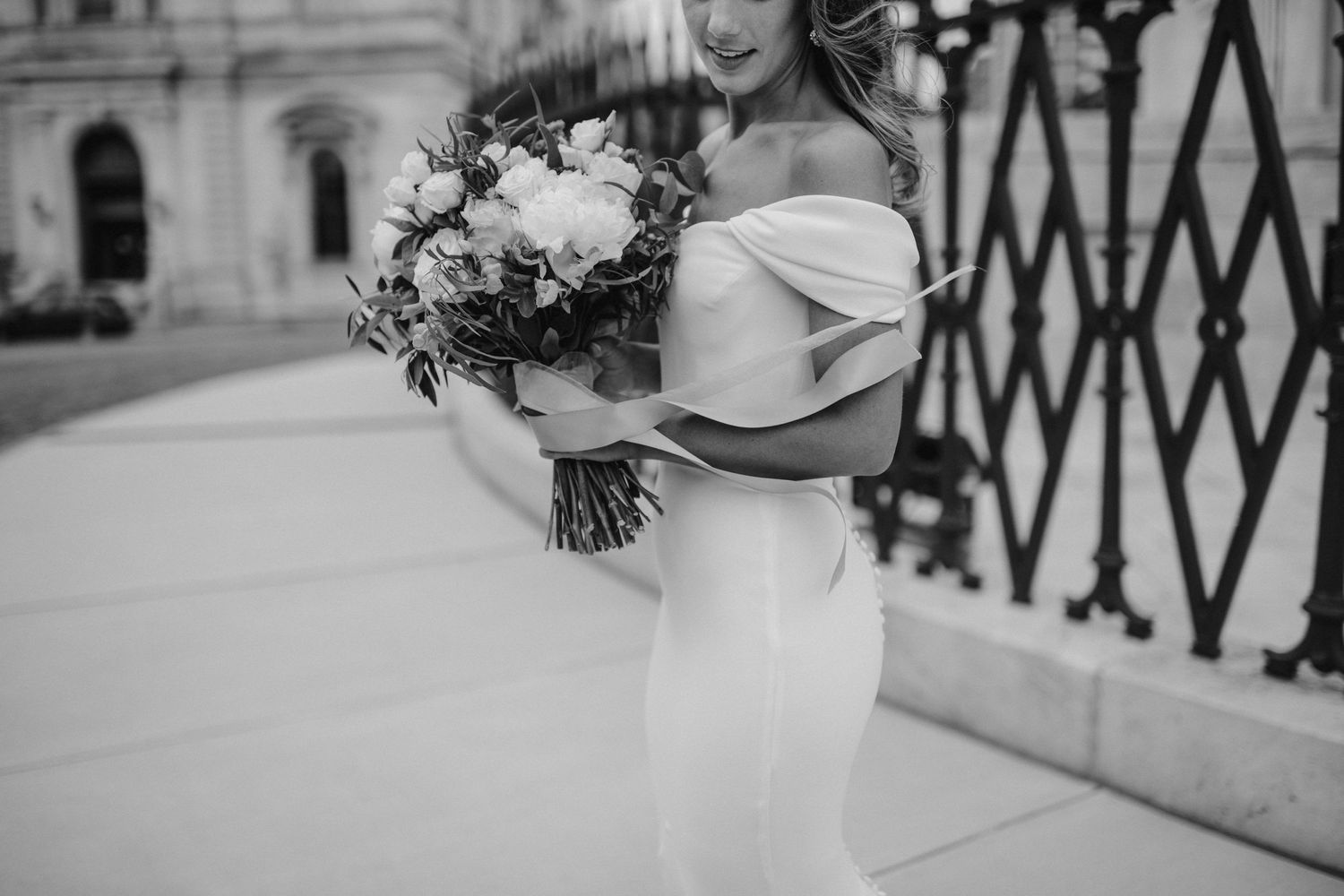 Black and white photo of a bride in a fitted off-shoulder wedding dress holding a lush bouquet.