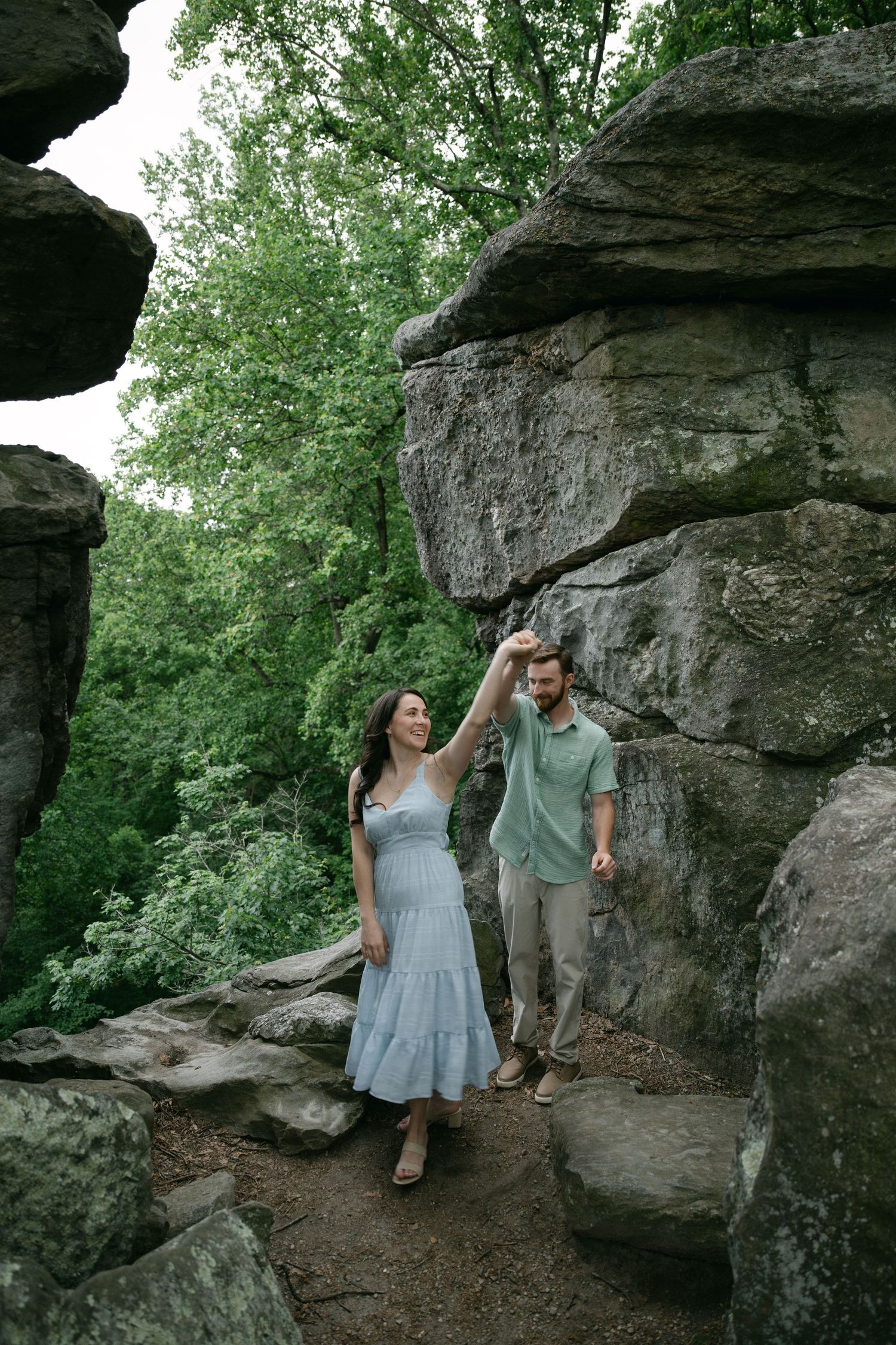 Two people share a tender moment while sitting on large rock formations in a wooded area.