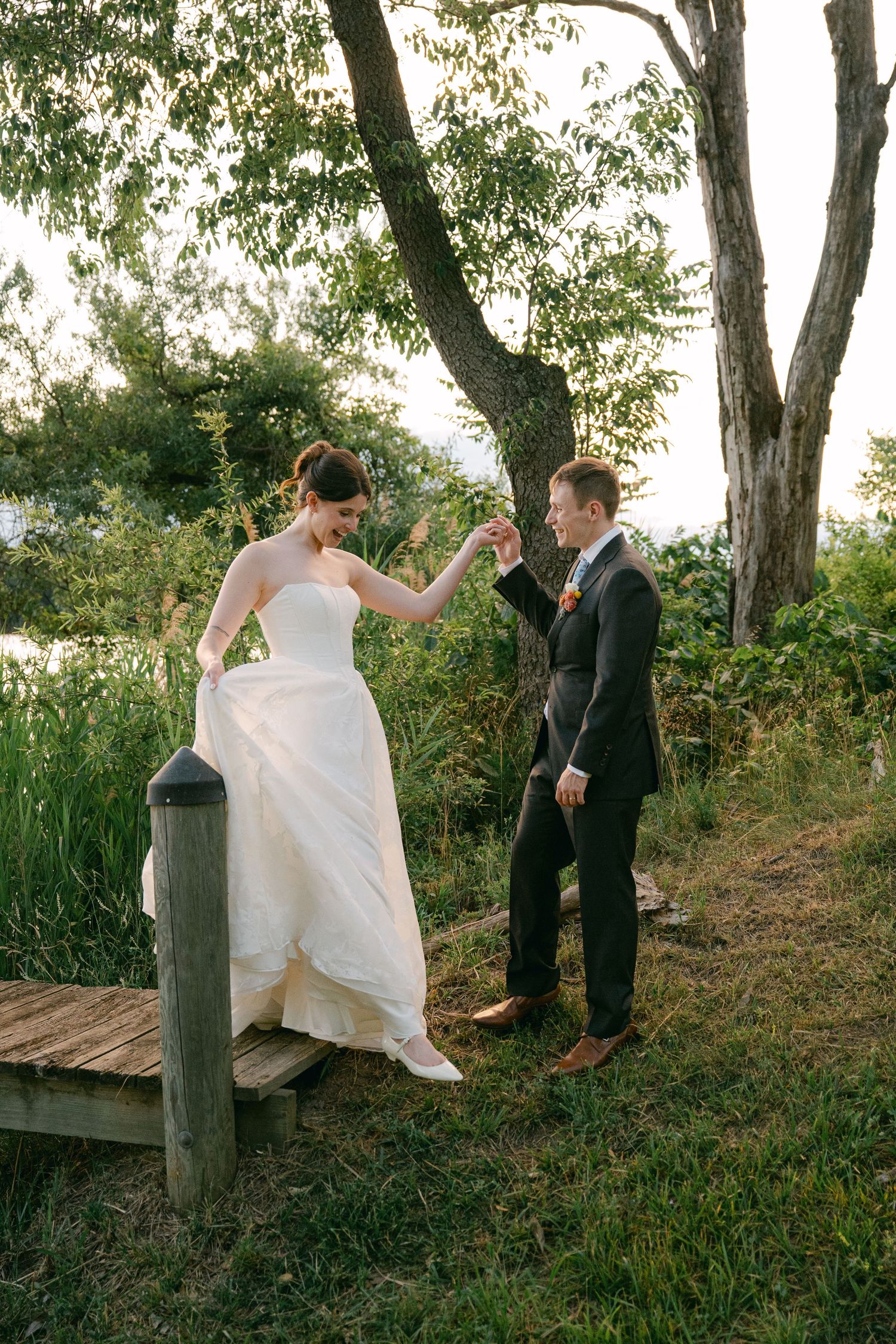 Groom helps bride step off wooden dock near pond, surrounded by lush greenery on their wedding day.
