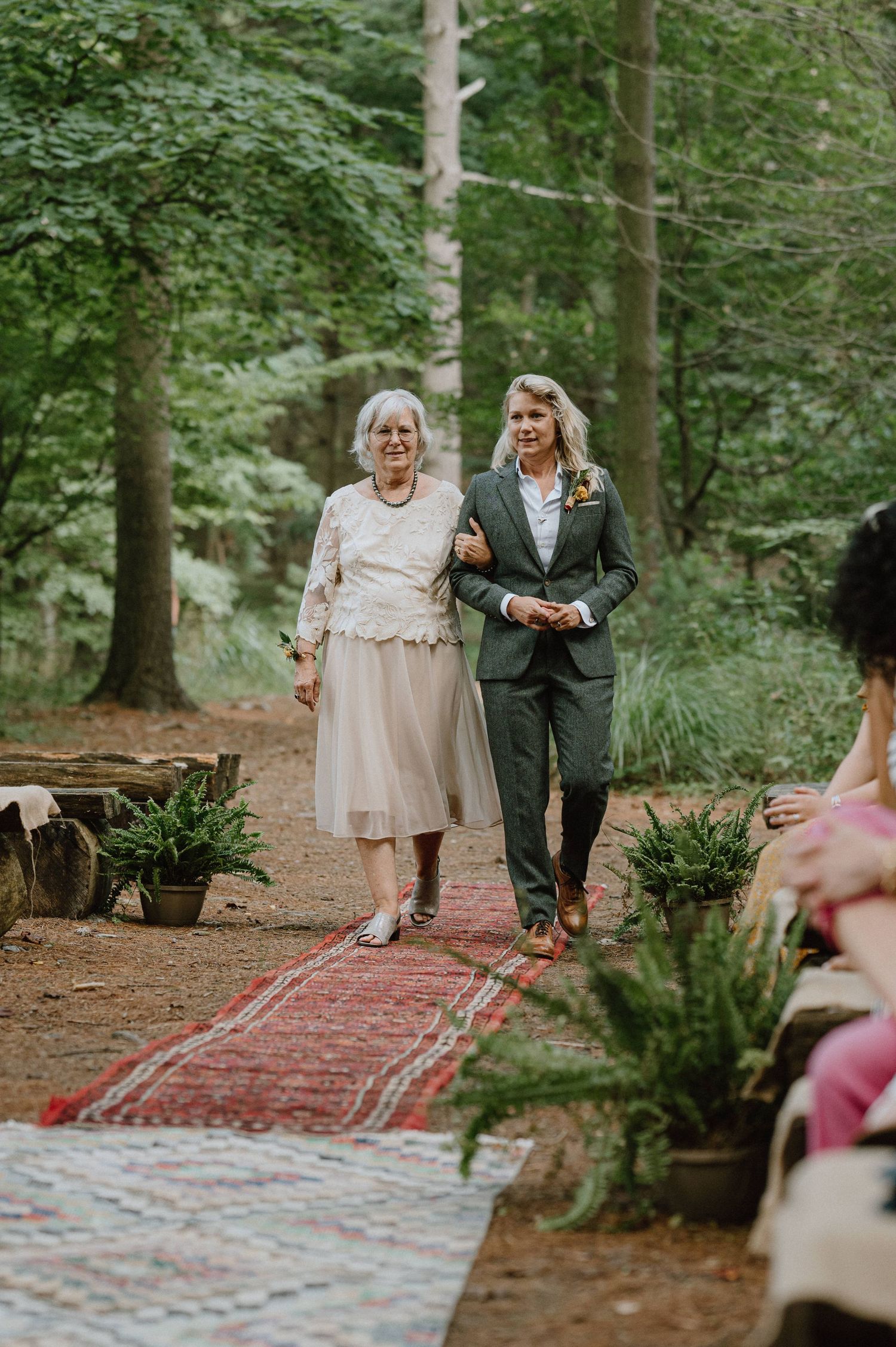 Two people walk down a red carpet aisle during an intimate forest wedding ceremony.