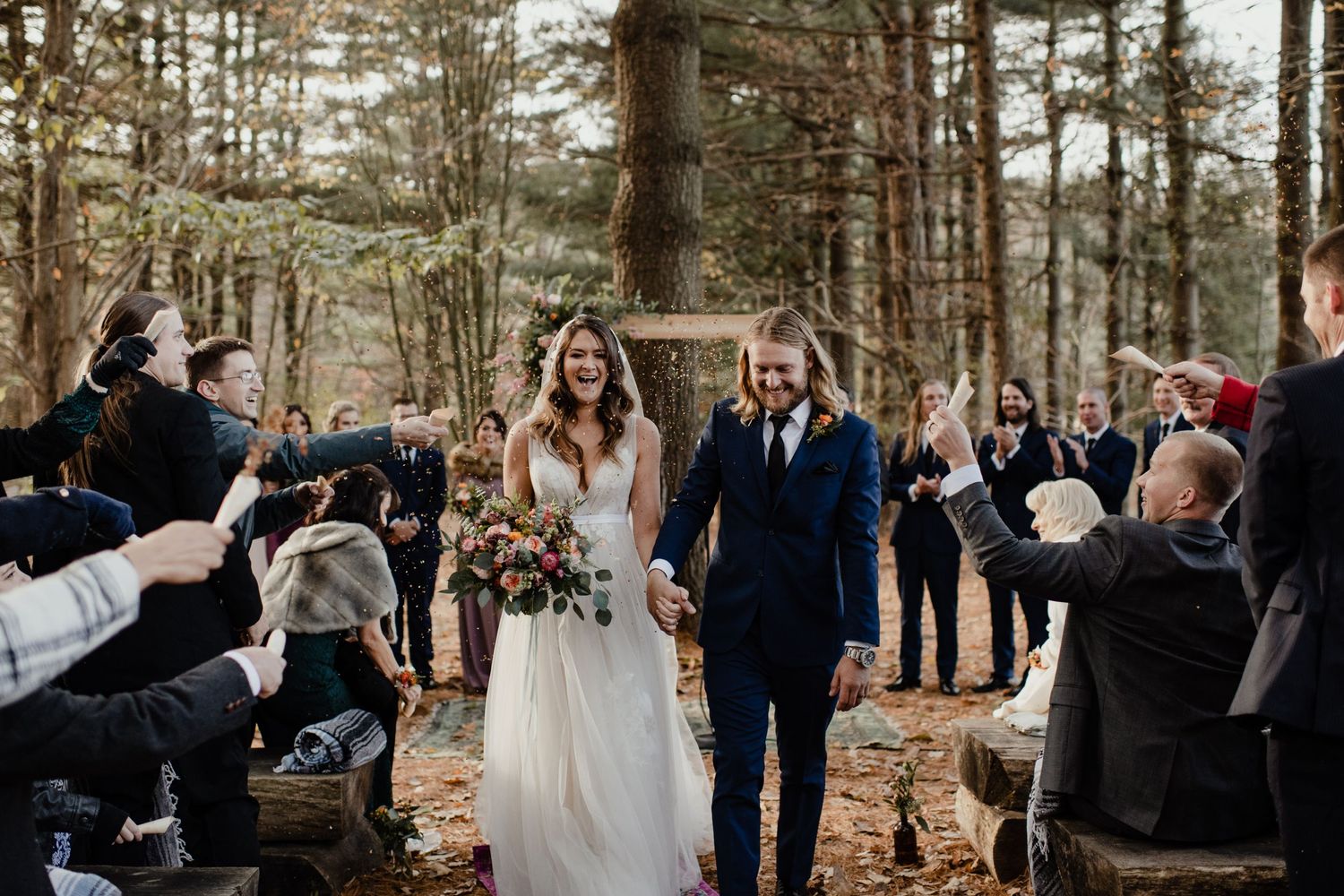 Guests cheer as newlyweds walk down a forest path during an outdoor celebration.