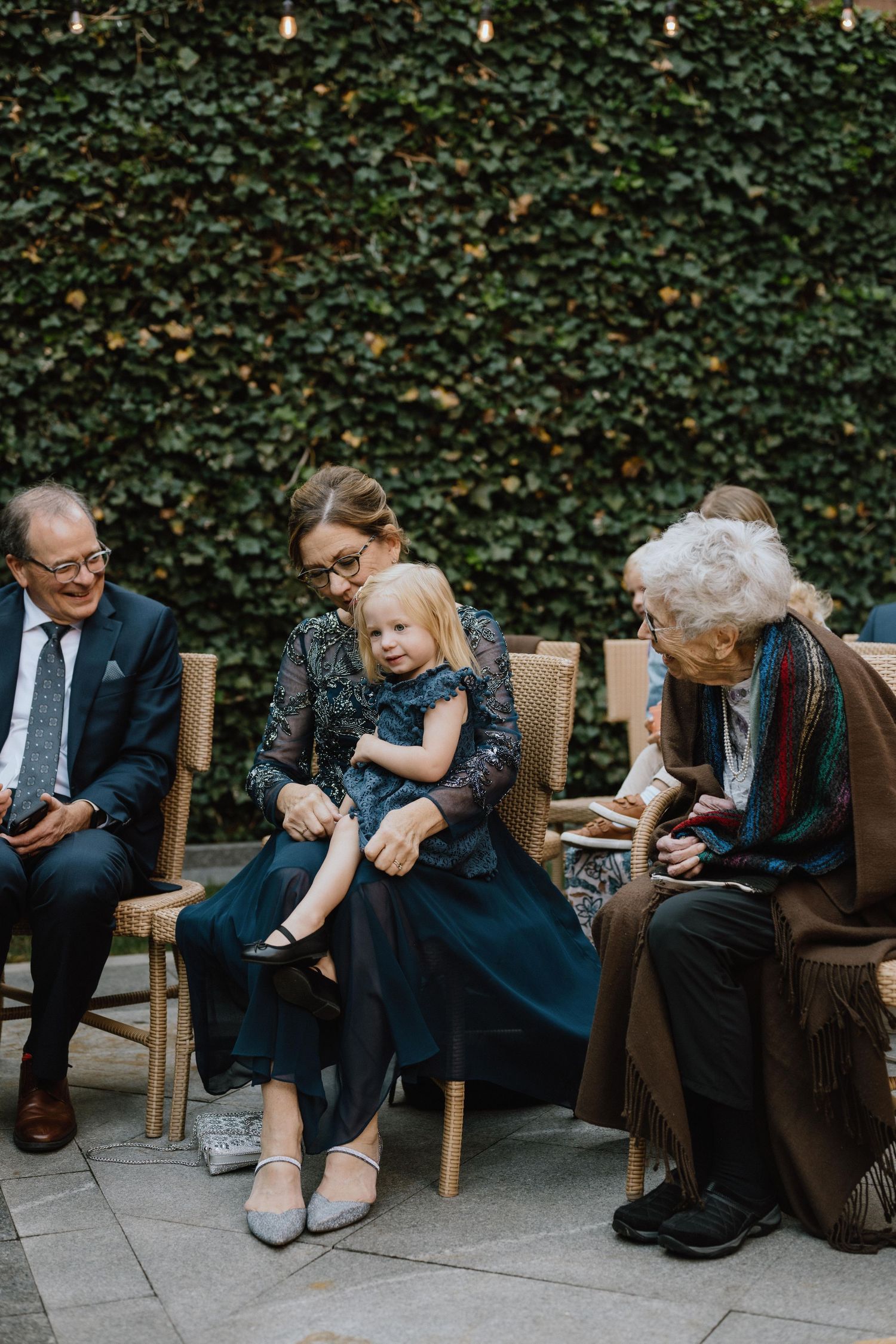 Multi-generational group seated on chairs against an ivy wall at an outdoor event.