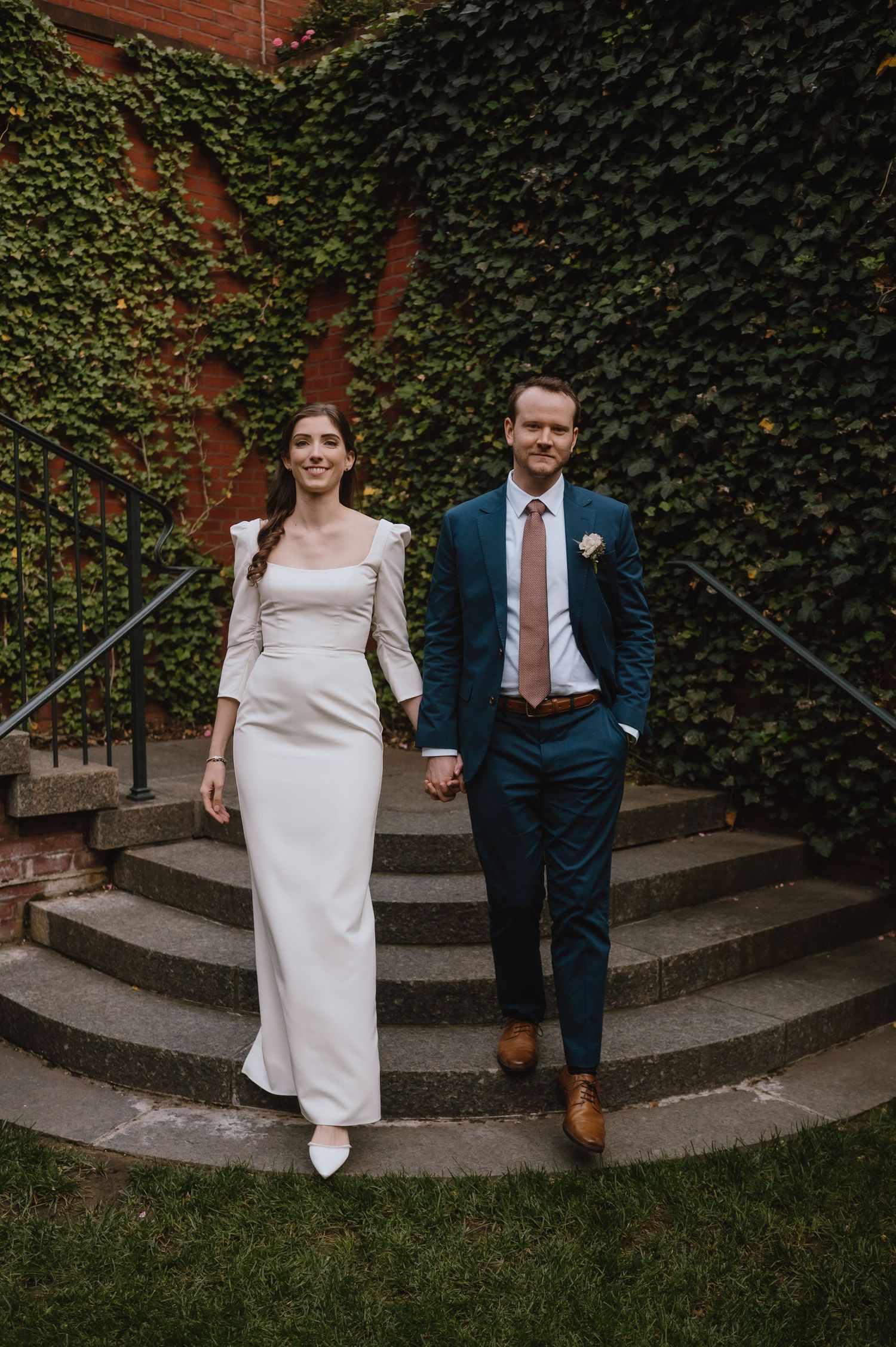 Elegant couple walking down stone steps with ivy-covered brick wall backdrop.